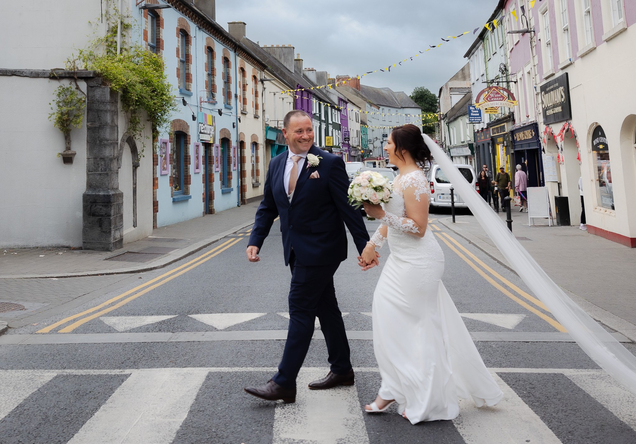 A newlywed couple holding hands and walking across a pedestrian crosswalk on a colorful urban street, with the groom wearing a dark suit and the bride in a white lace wedding gown, holding a bouquet of flowers.