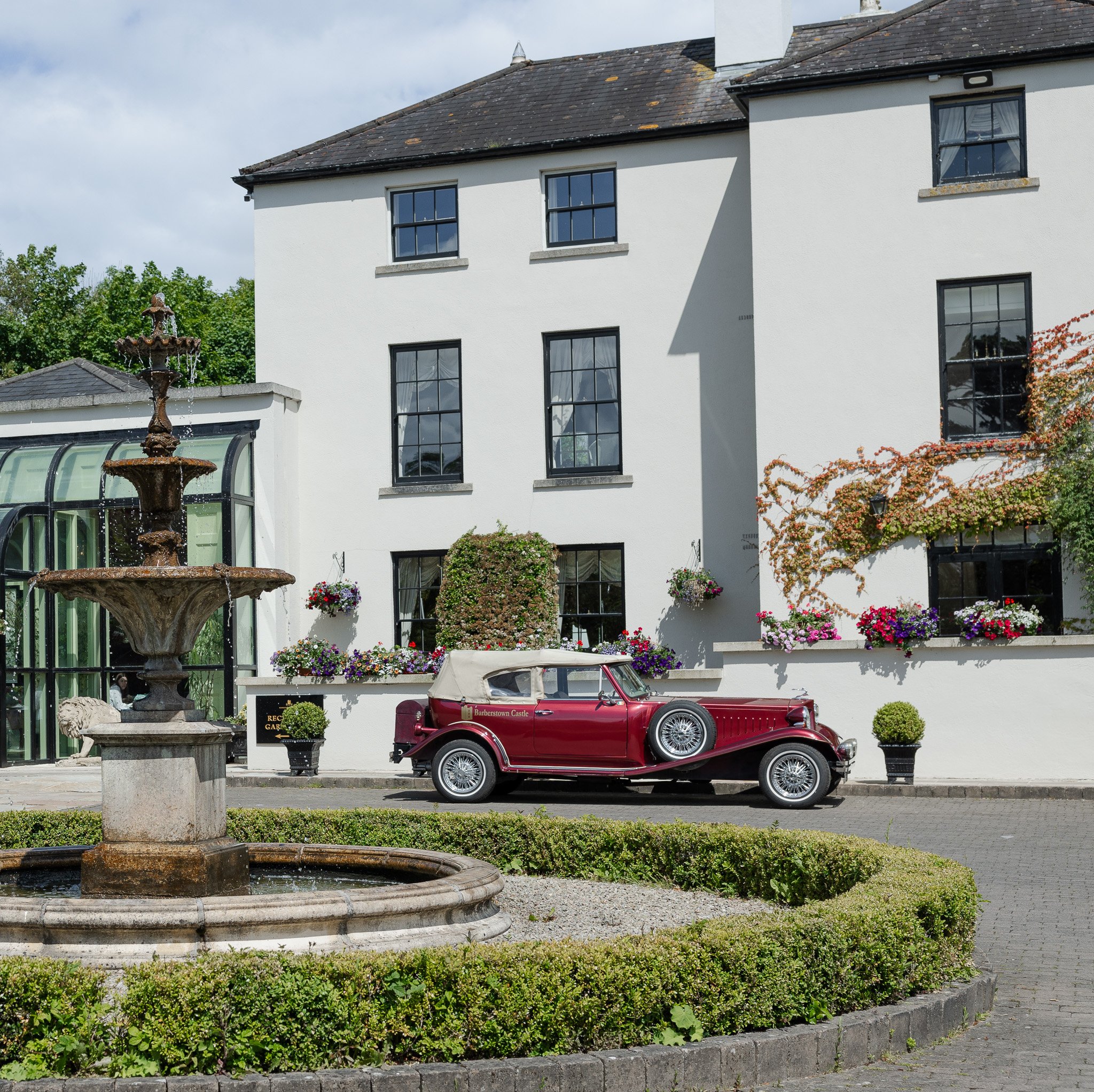 A vintage red convertible car parked in front of a white building with multiple windows, surrounded by colorful flowers and a water fountain.