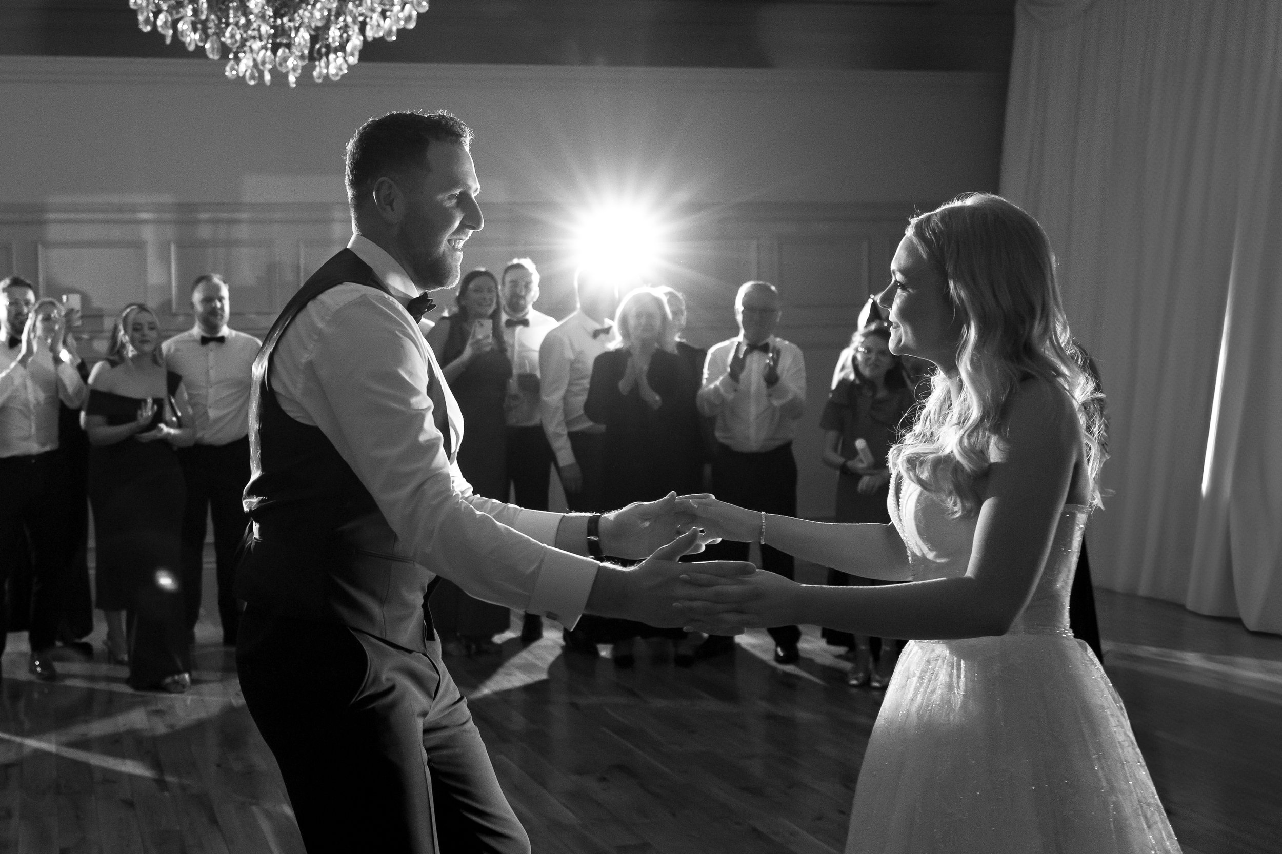 Black and white photo of a bride and groom dancing at their wedding reception, surrounded by guests watching and smiling.