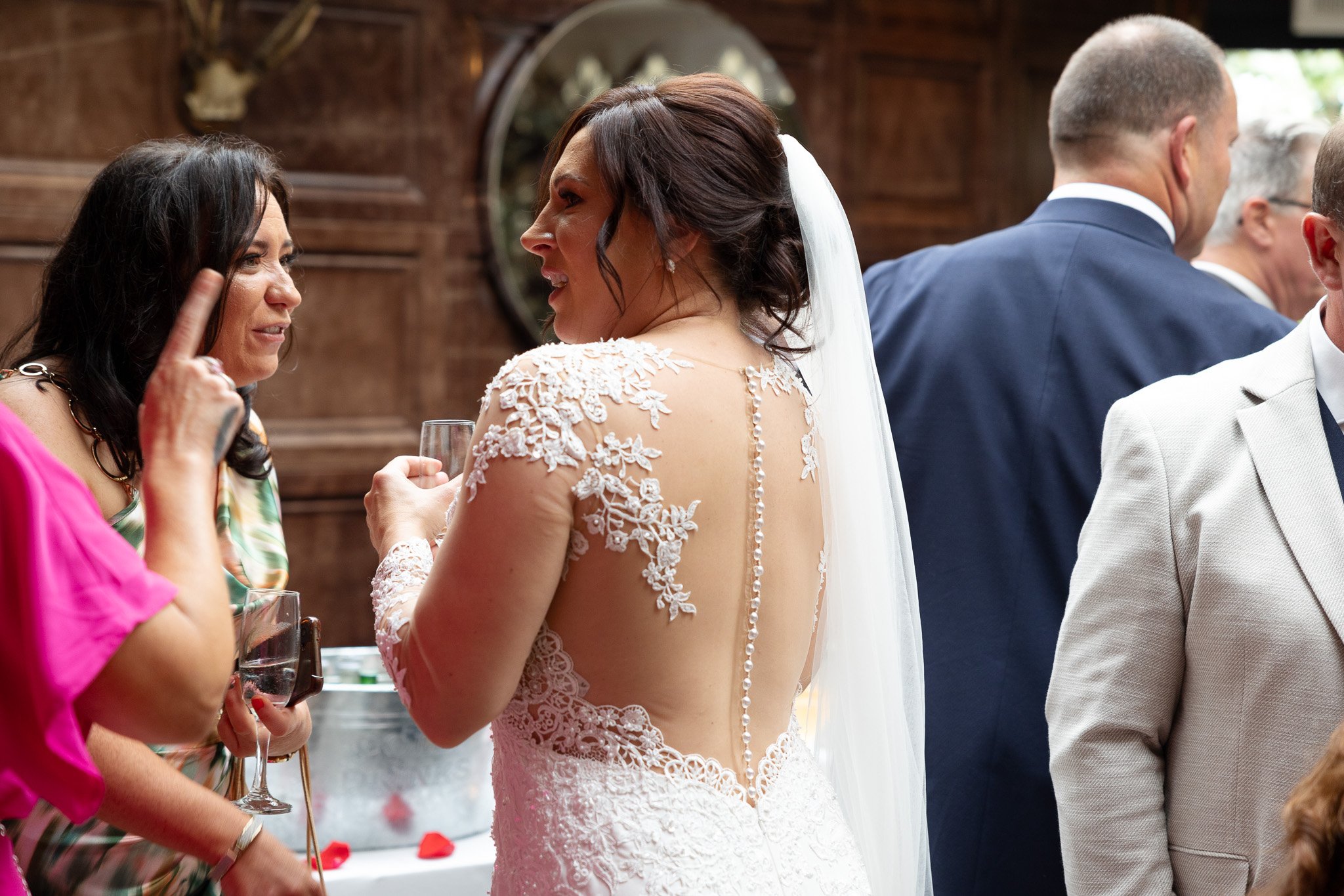 A bride and a woman engaged in conversation at a wedding reception. The bride is wearing a lace wedding dress with an open back and veil, holding a glass of wine. The woman is wearing a colorful patterned dress and has long dark hair. Several other g