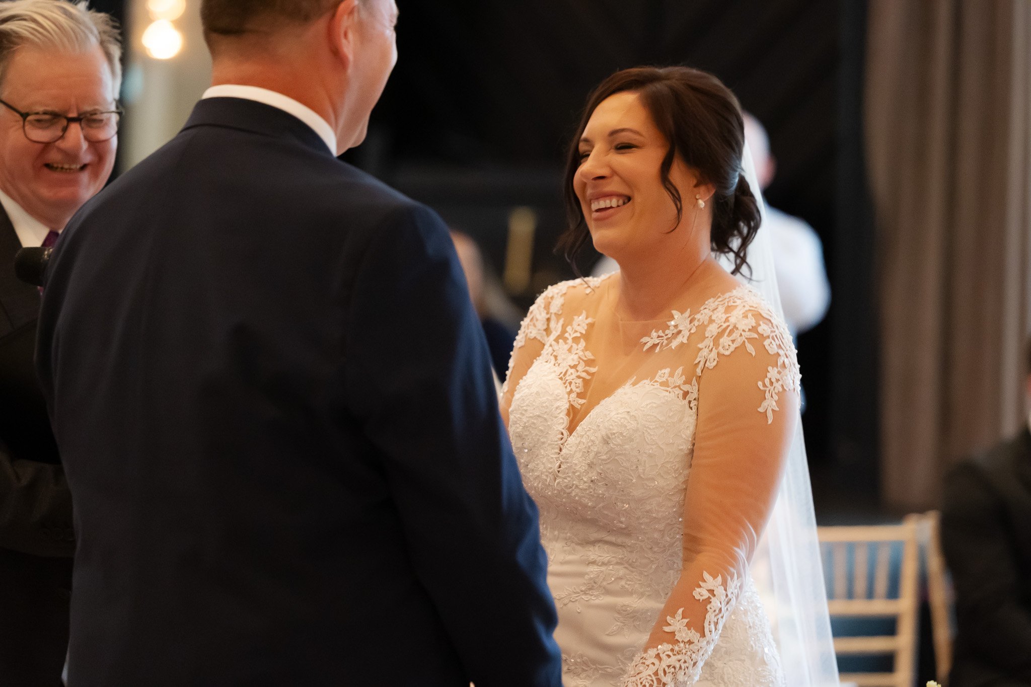 A bride smiling during her wedding ceremony, standing with the officiant and groom in a formal setting.