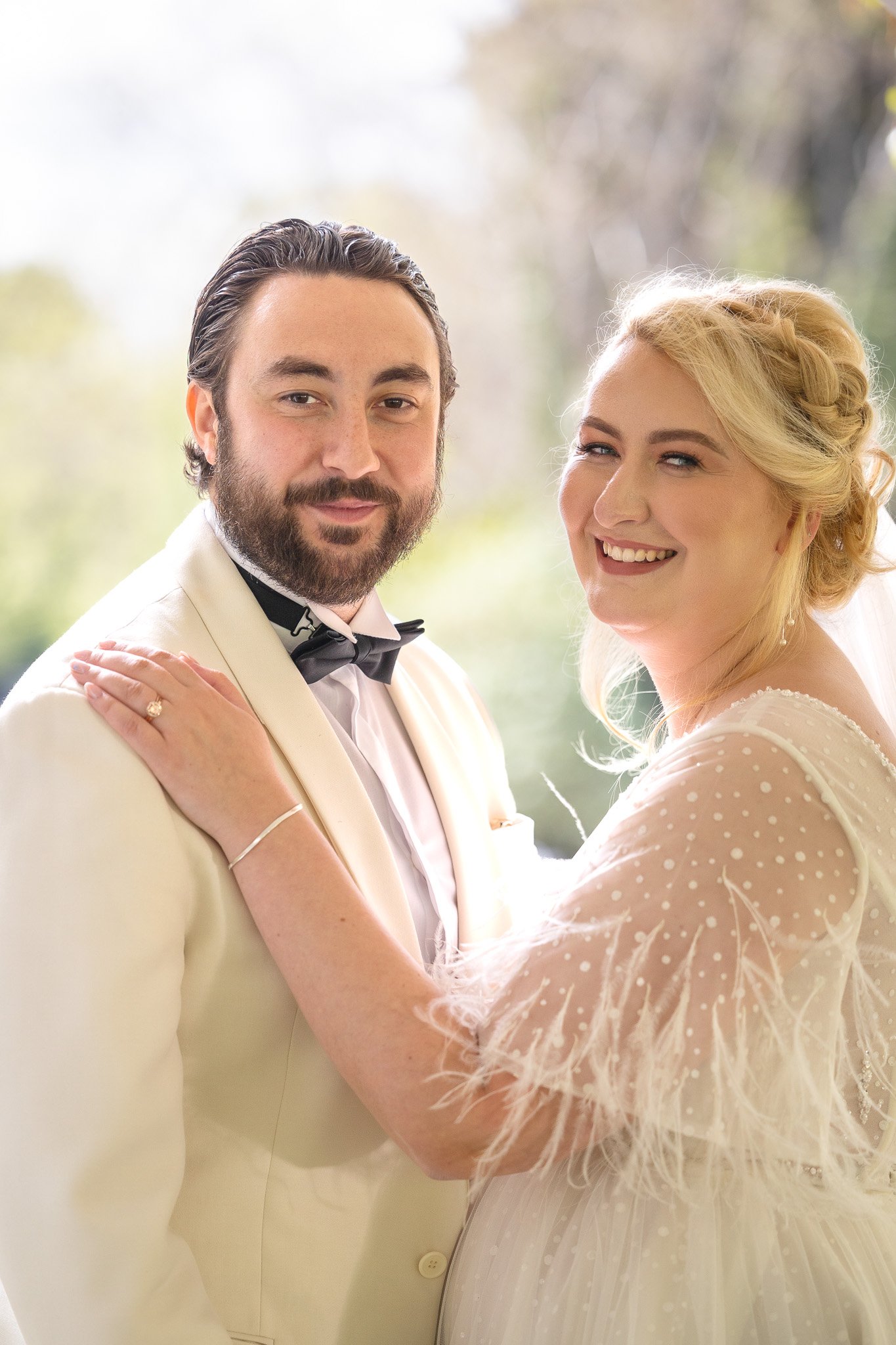 A bride and groom on their wedding day, smiling and embracing outdoors with a blurred natural background.