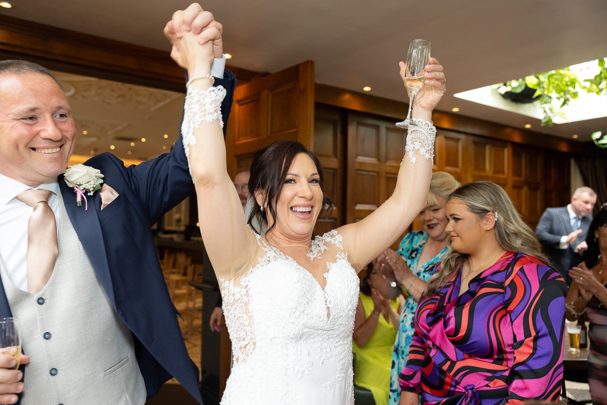 Bride and groom celebrating at a wedding reception, with arms raised and holding champagne glasses, surrounded by guests.