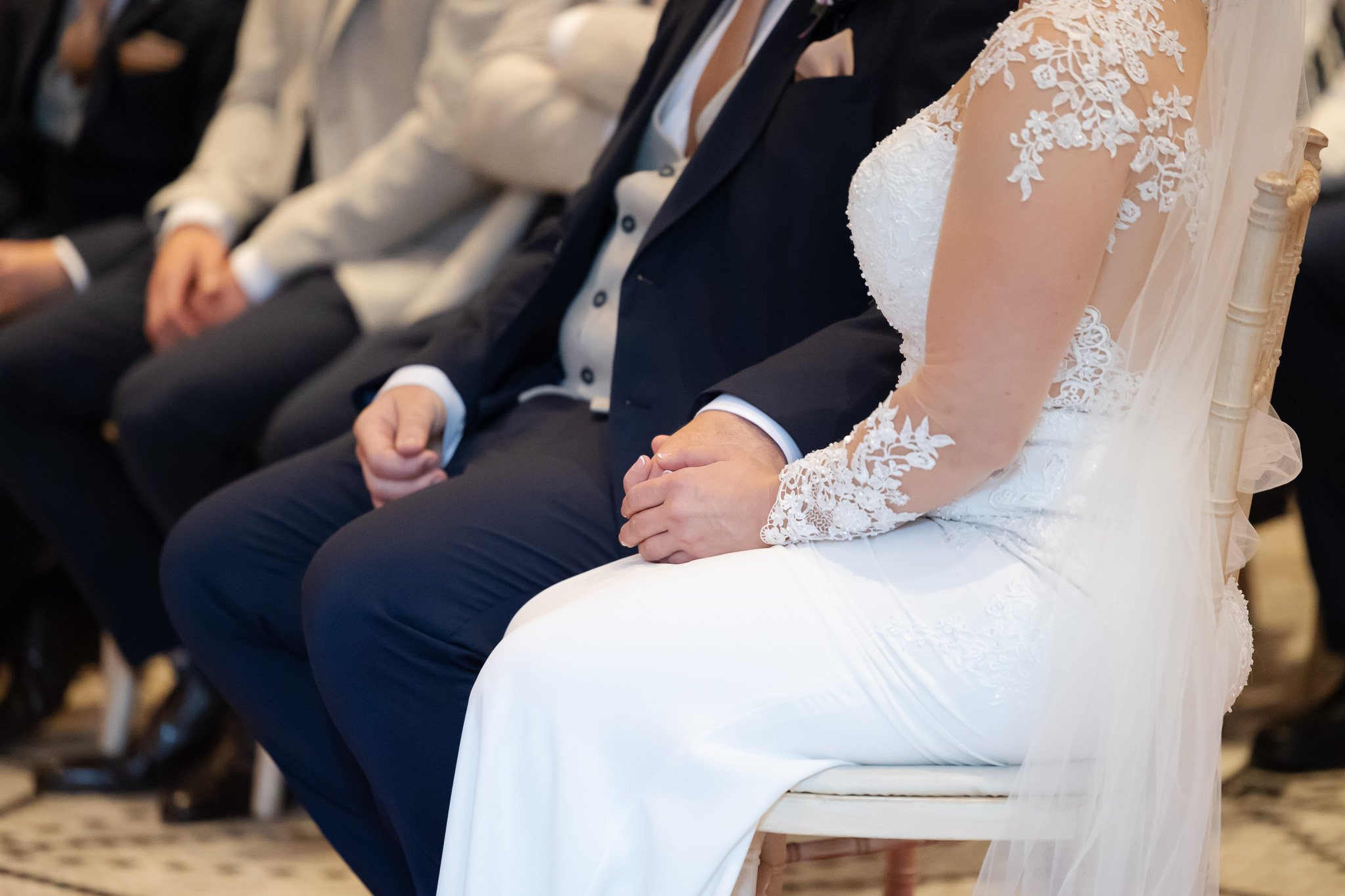 Close-up of a bride and groom holding hands during a wedding ceremony, seated on chairs in front of a seated audience.