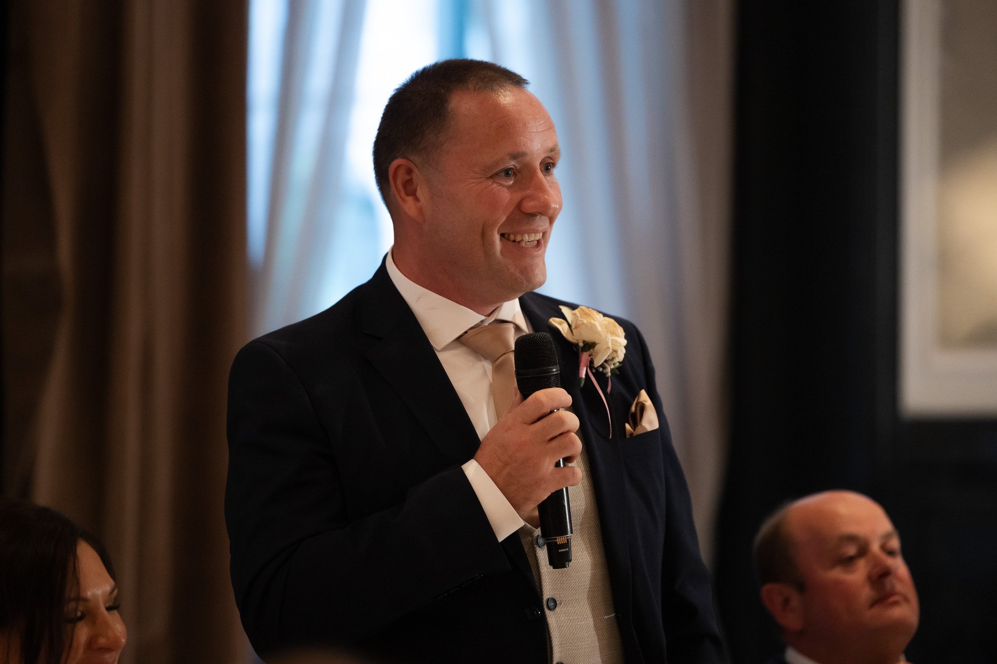 A man in a dark suit and beige tie holding a microphone, smiling, at a formal event or wedding reception.