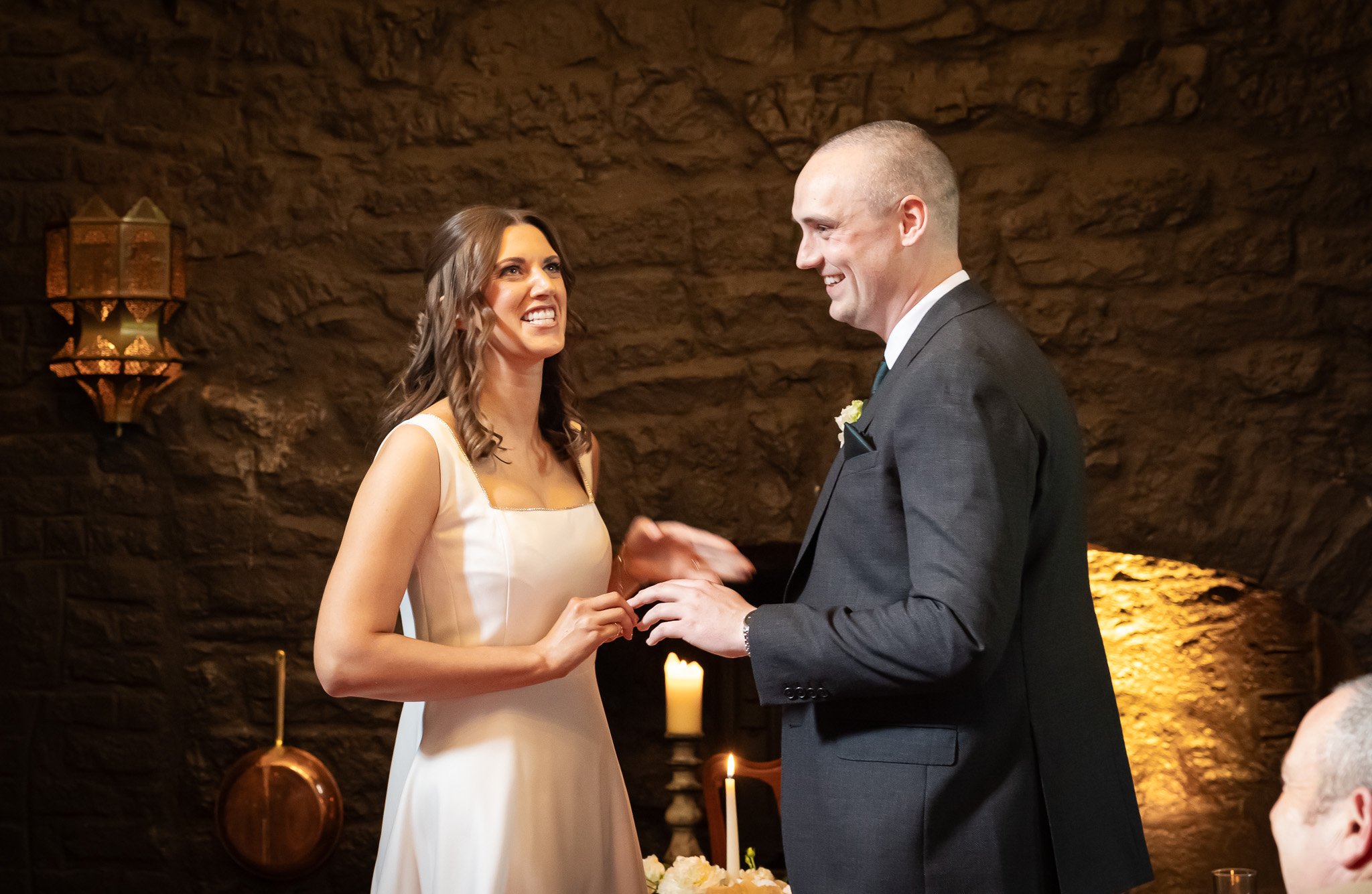 A bride and groom exchange vows or rings during their wedding ceremony in front of a rustic brick wall with hanging lanterns and candles.