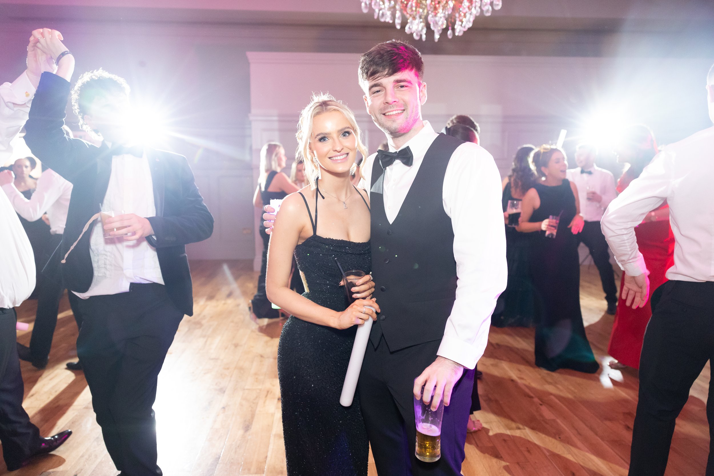 Couple at a formal dance or event, smiling and dressed in tuxedo and gown, holding drinks among others dancing in the background, with bright lights and chandelier overhead.