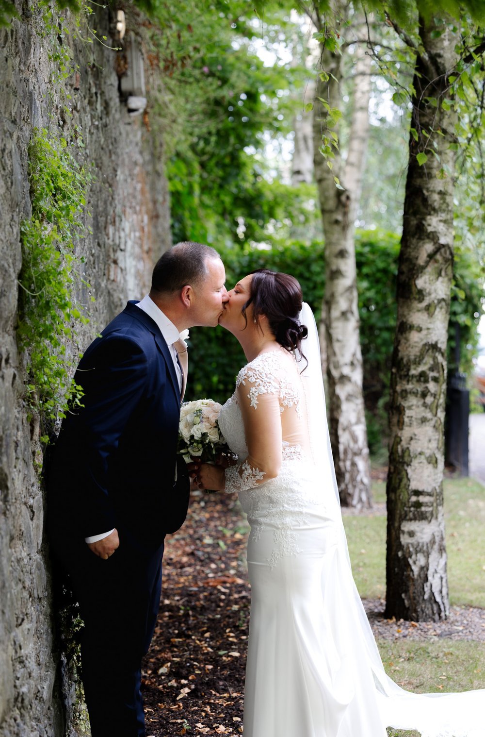 A bride and groom share a kiss outdoors next to a stone wall and trees, with the bride holding a bouquet of white flowers.