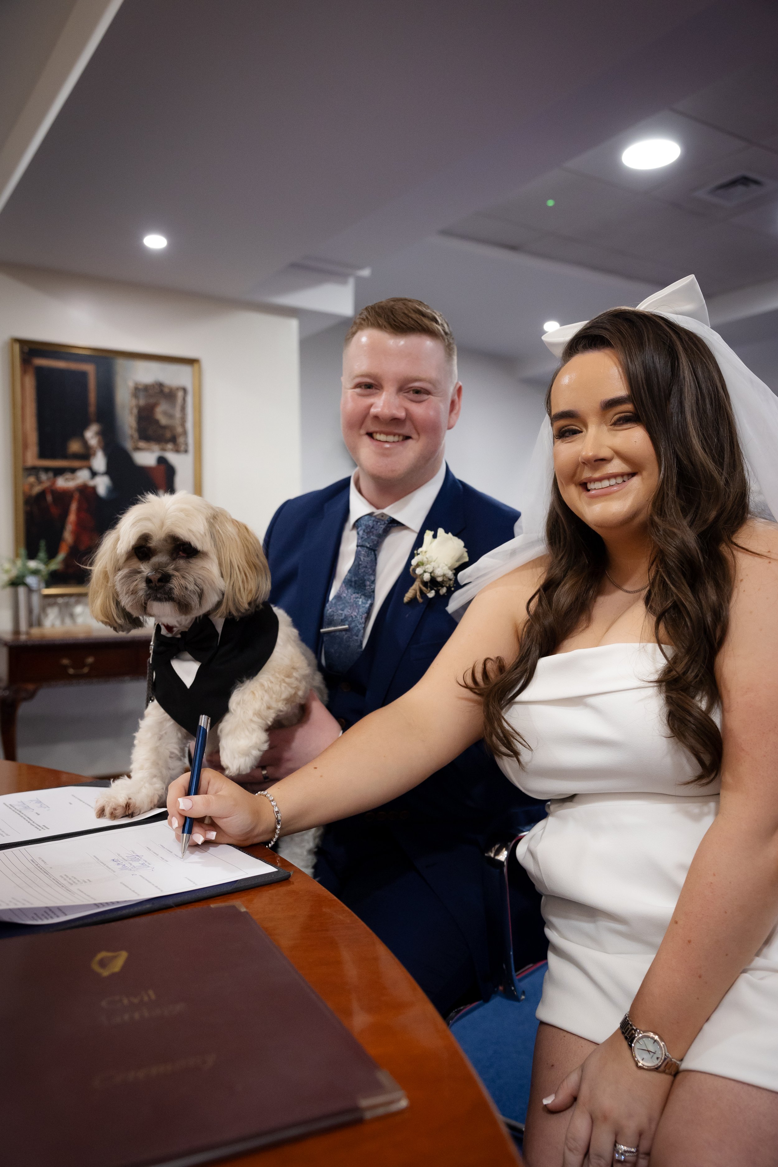 A smiling couple signing a marriage certificate at a wedding ceremony, with a small dog dressed in a tuxedo on the table in front of them.
