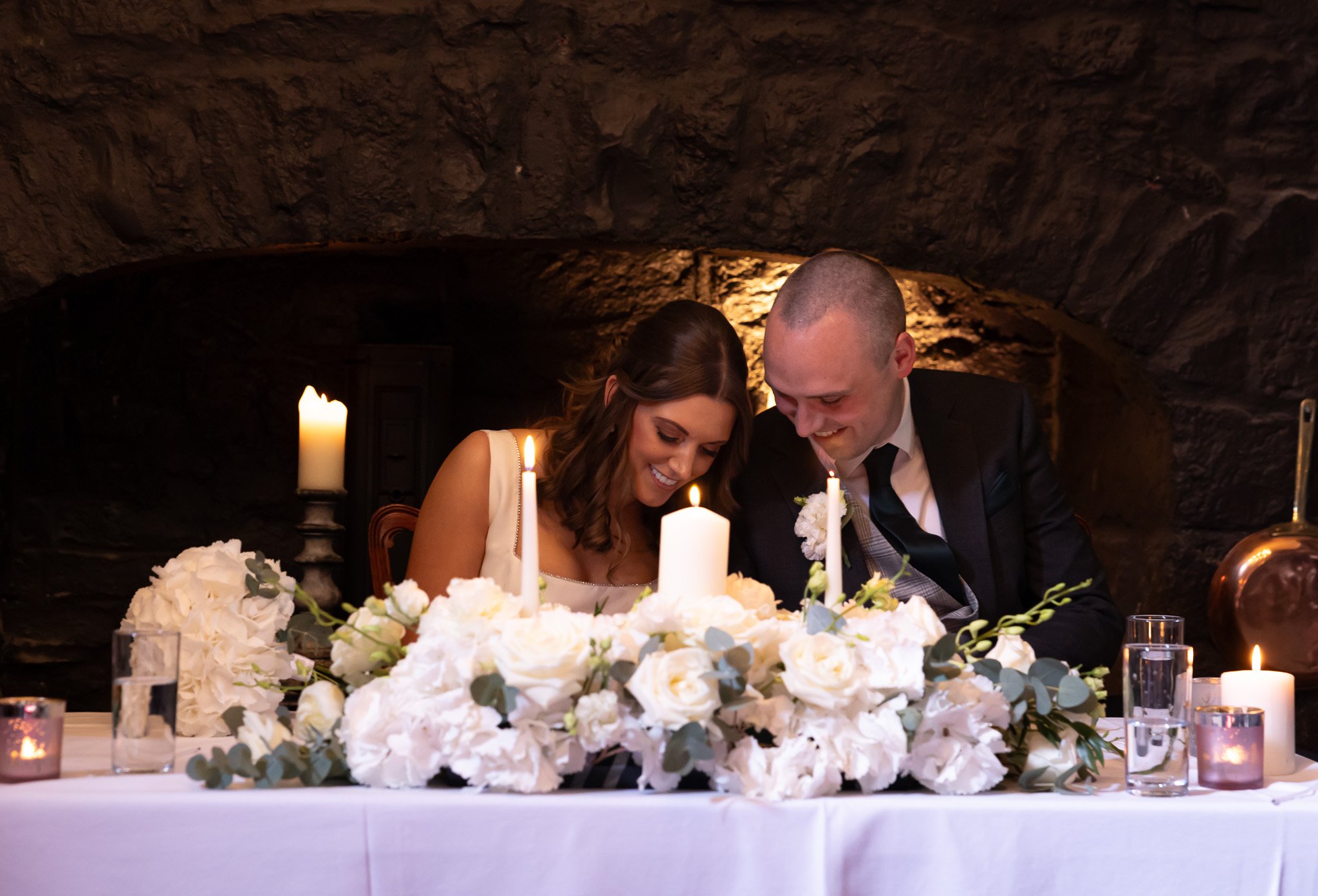 A wedding couple, a bride and groom, smiling and leaning close together at a table decorated with white flowers and candles, in a dimly lit setting with a brick or stone background.