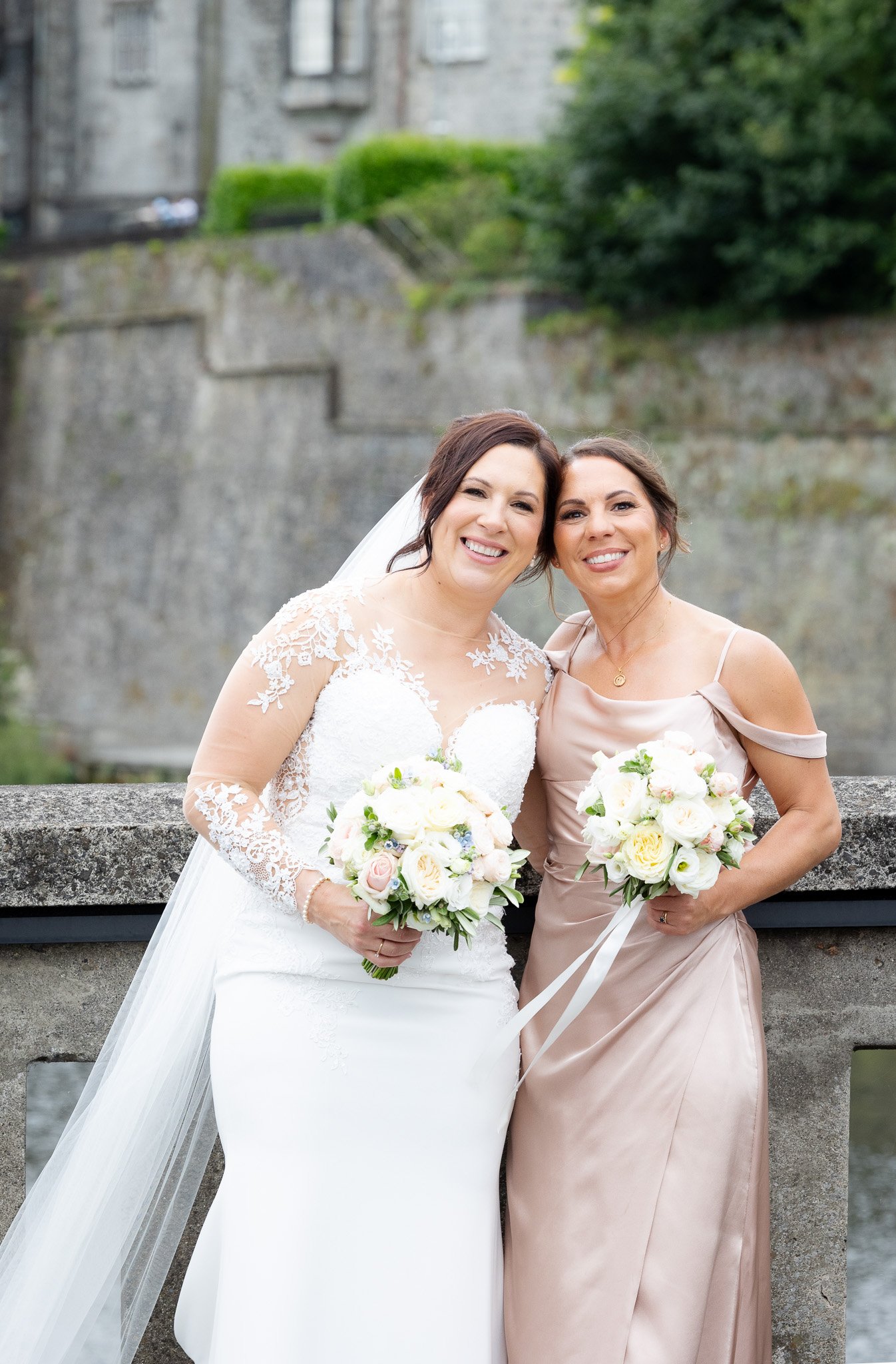 Two women, one in a wedding dress and the other in a pastel gown, standing on a bridge, holding bouquets, smiling.