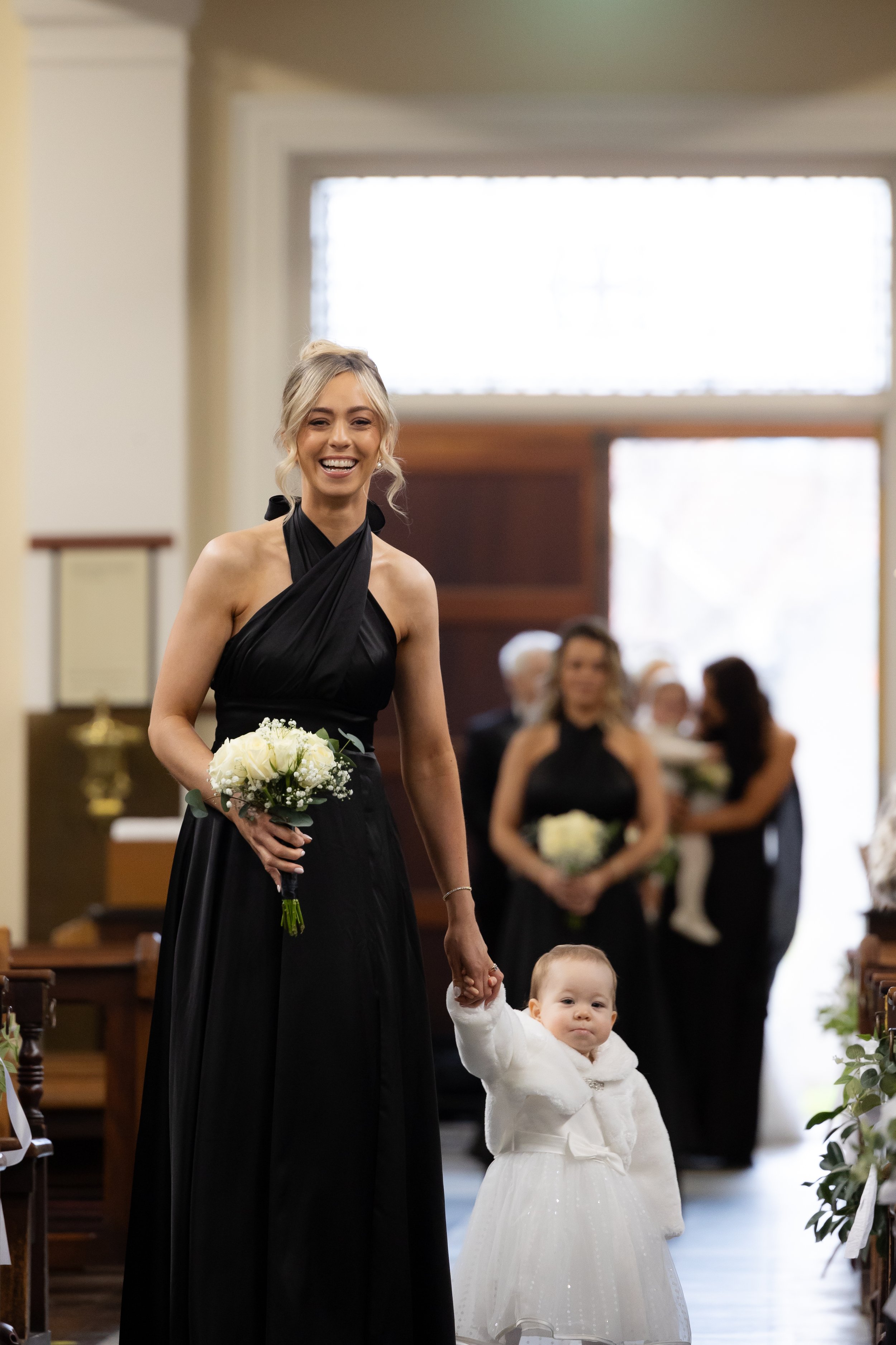 A woman in a black dress holding a bouquet of white flowers and a child's hand, smiling in a church during a wedding ceremony.