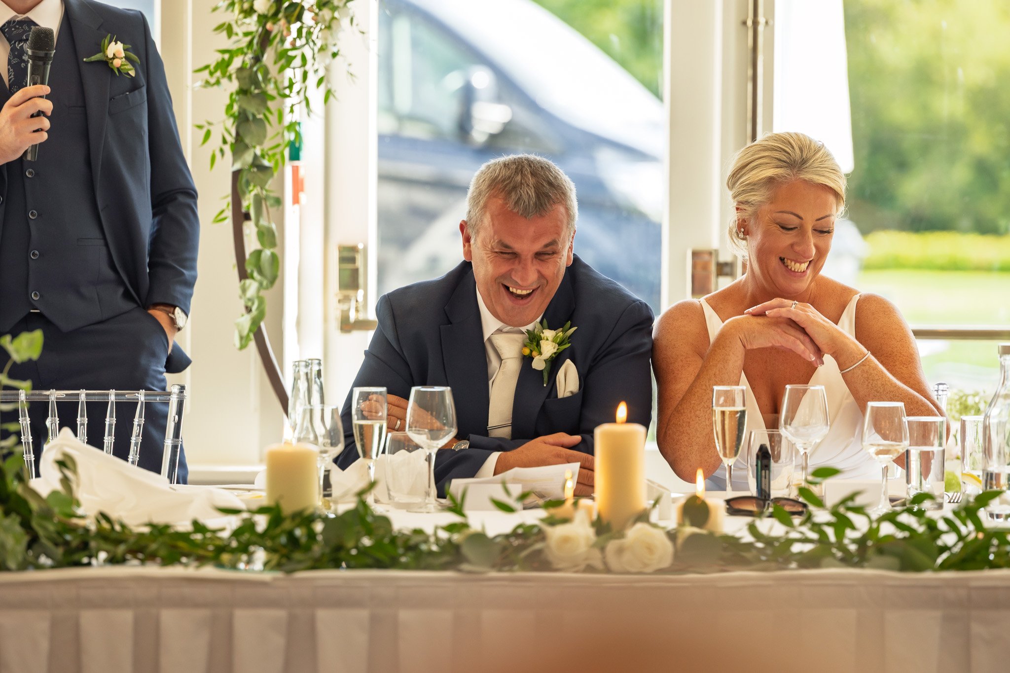 A bride and groom sitting at a decorated table during their wedding reception, smiling and laughing. The table has candles, glasses, and floral arrangements. A man in a dark suit is standing nearby with a microphone.