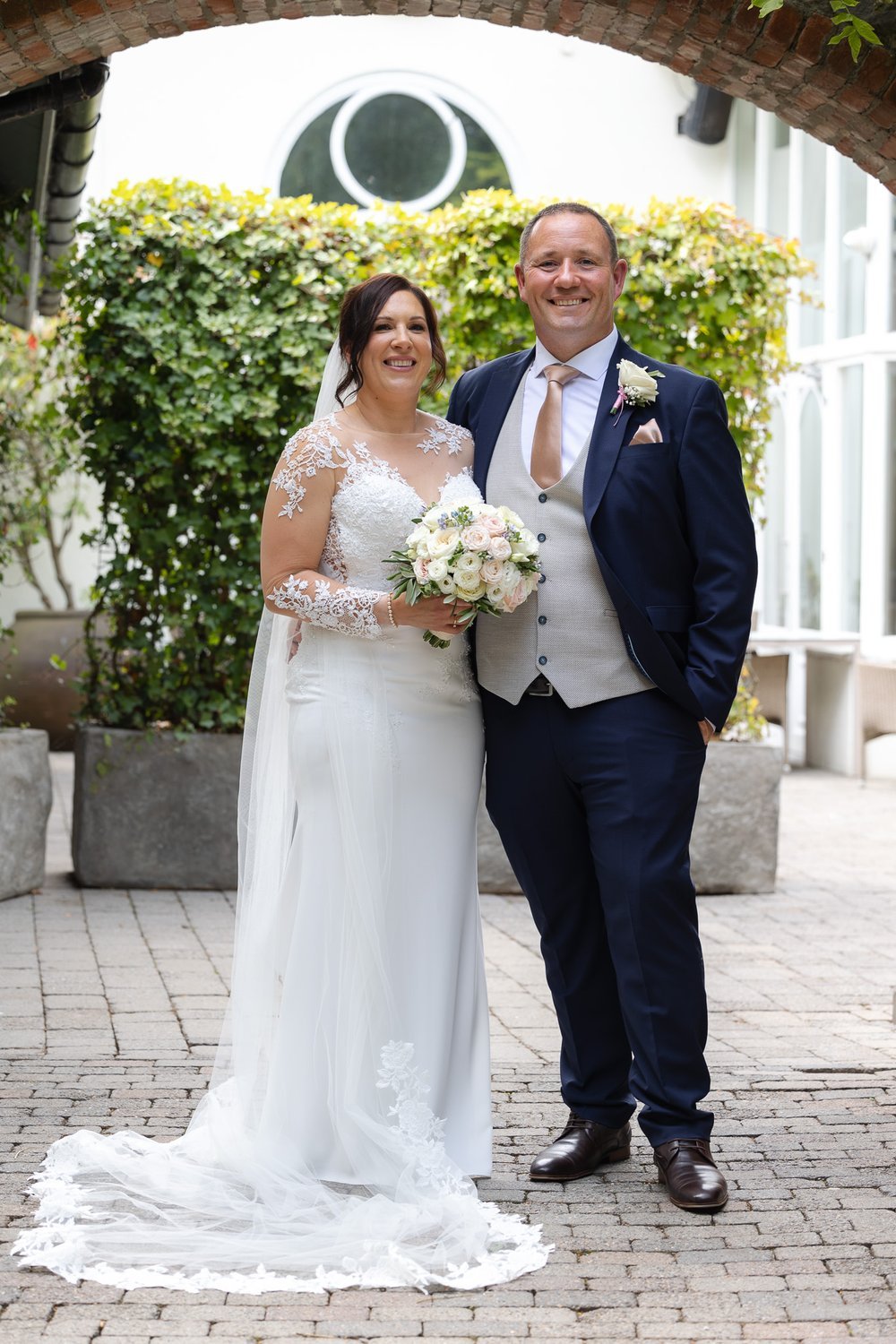 A bride and groom pose for a wedding photo outdoors under an archway. The bride is in a white lace wedding dress holding a bouquet of white and pink roses. The groom is in a navy suit with a beige vest, pink tie, and boutonniere. Green shrubbery and 