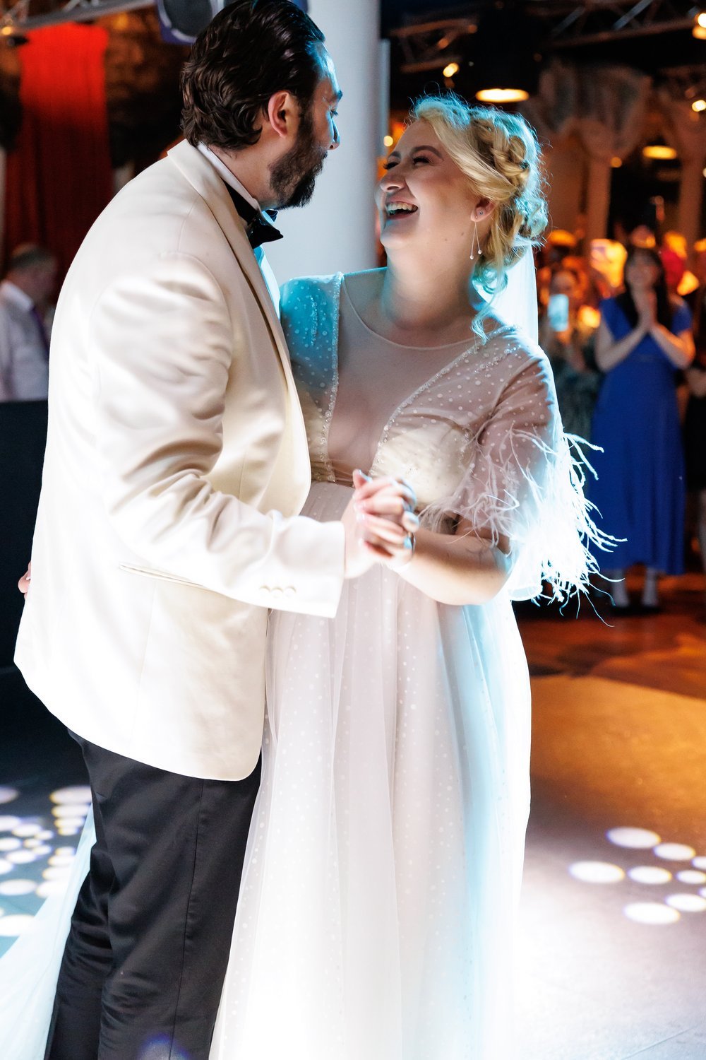 A smiling couple dancing at their wedding reception, the man wearing a white tuxedo jacket and the woman in a white wedding gown with feathered sleeves, surrounded by joyful guests in a warmly lit venue.