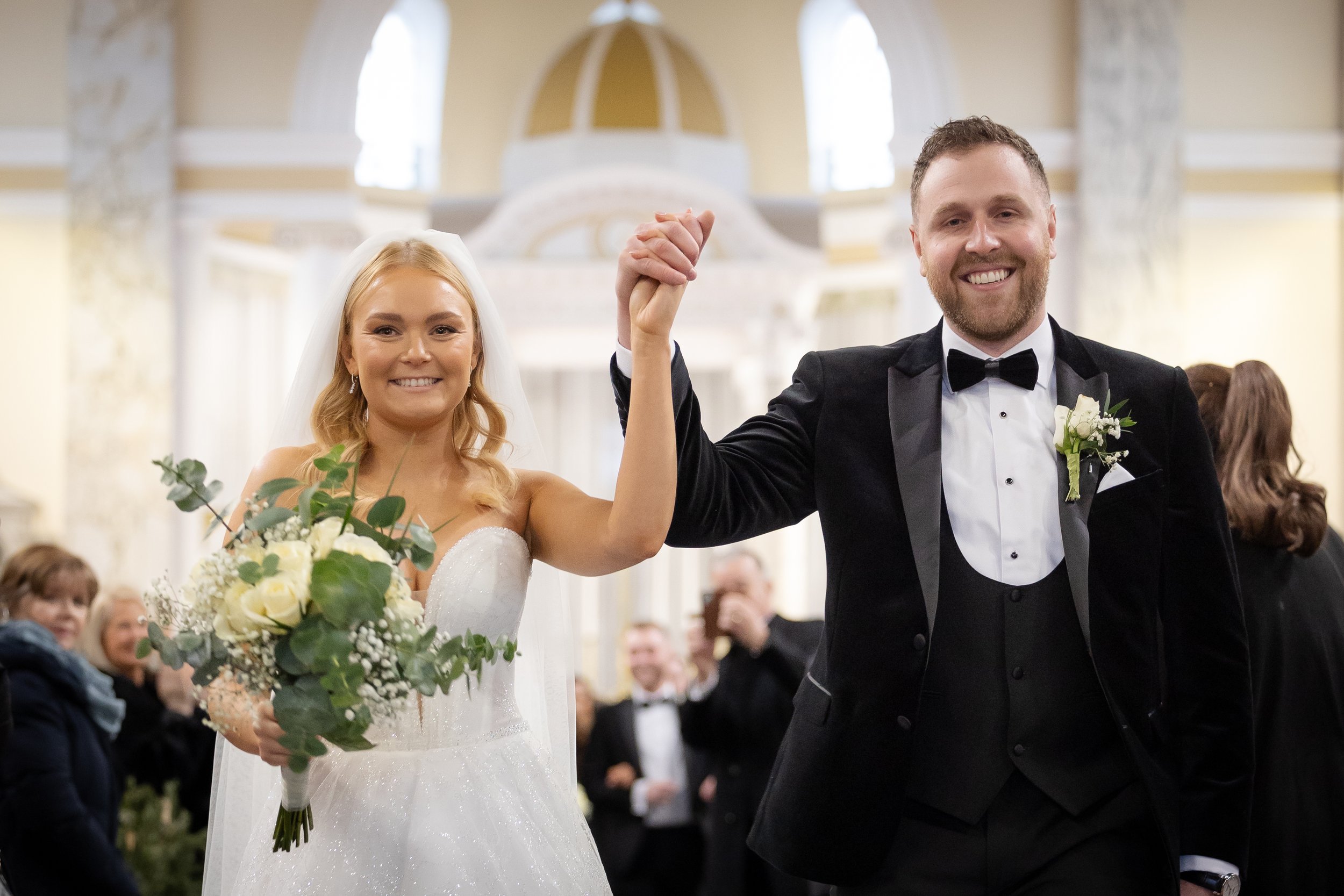 Happy bride and groom celebrating wedding, holding hands with arms raised, in a decorated church with guests in the background.
