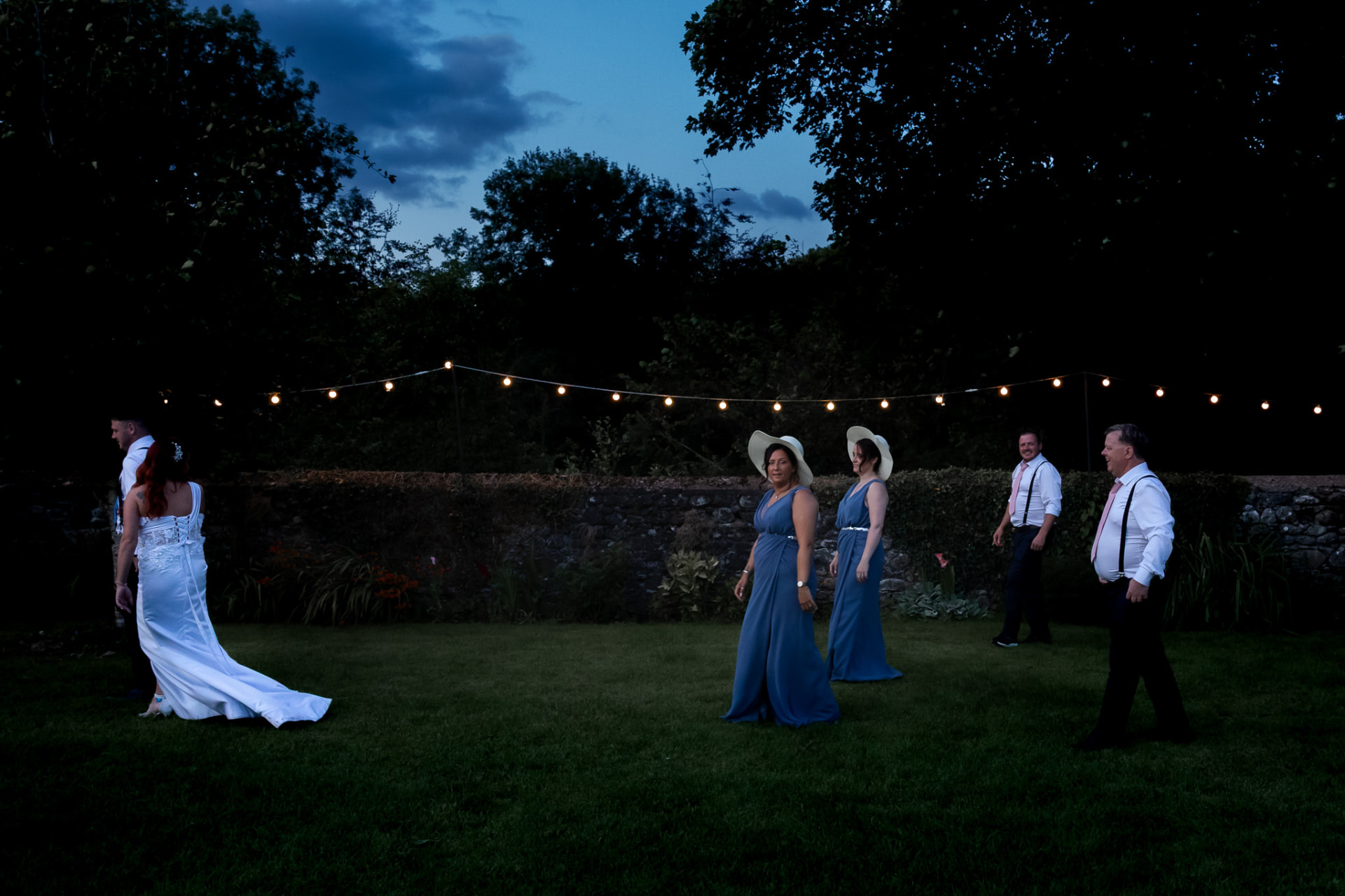 Wedding party gathered on the lawn beneath festoon lights at dusk