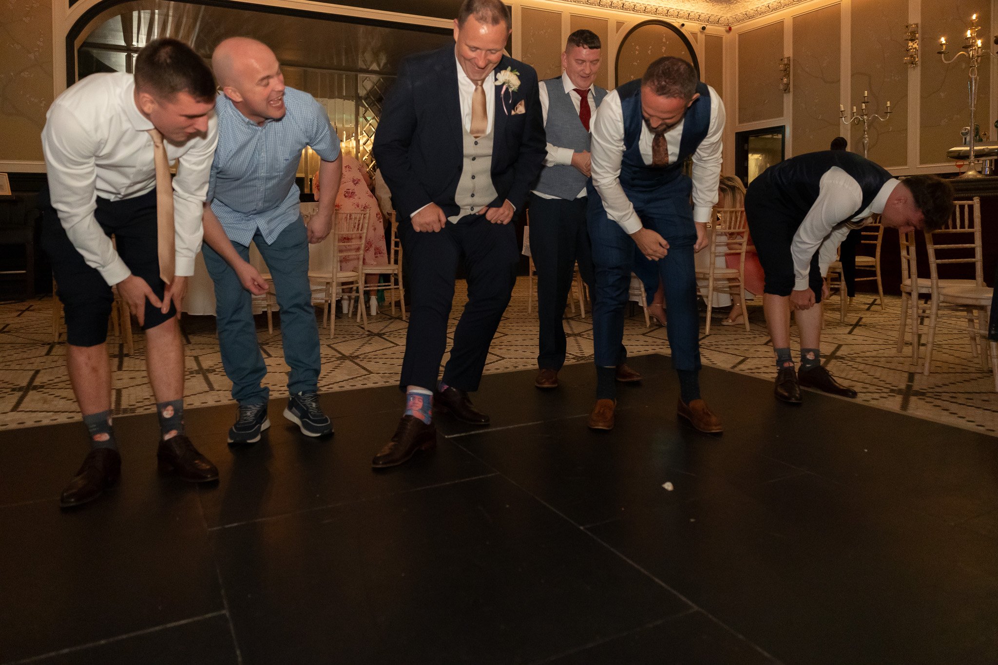 Six men in formal attire engaged in a limbo dance at an indoor event, bending backwards under a horizontal bar, with a decorated banquet hall background.