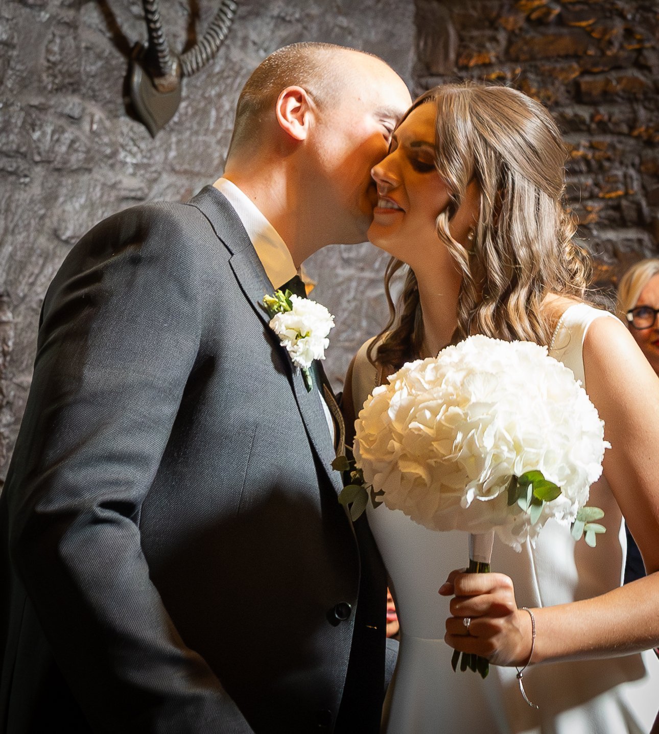 A bride and groom sharing a kiss at their wedding ceremony, with the bride holding a bouquet of white flowers and wearing a white dress, and the groom in a dark suit with a boutonnière, standing against a stone wall background.