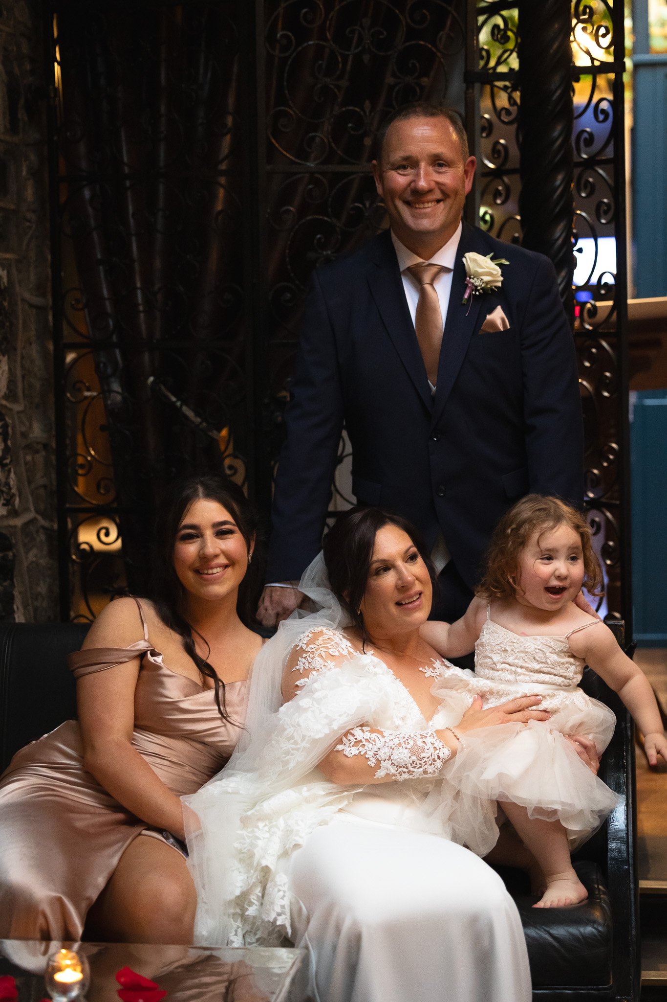 A group of four people, two women, one man, and a young girl, gathered together at a wedding celebration; the man is in a dark suit with a boutonniere, the women are in elegant dresses, and the girl is in a light dress. They are smiling and posing fo