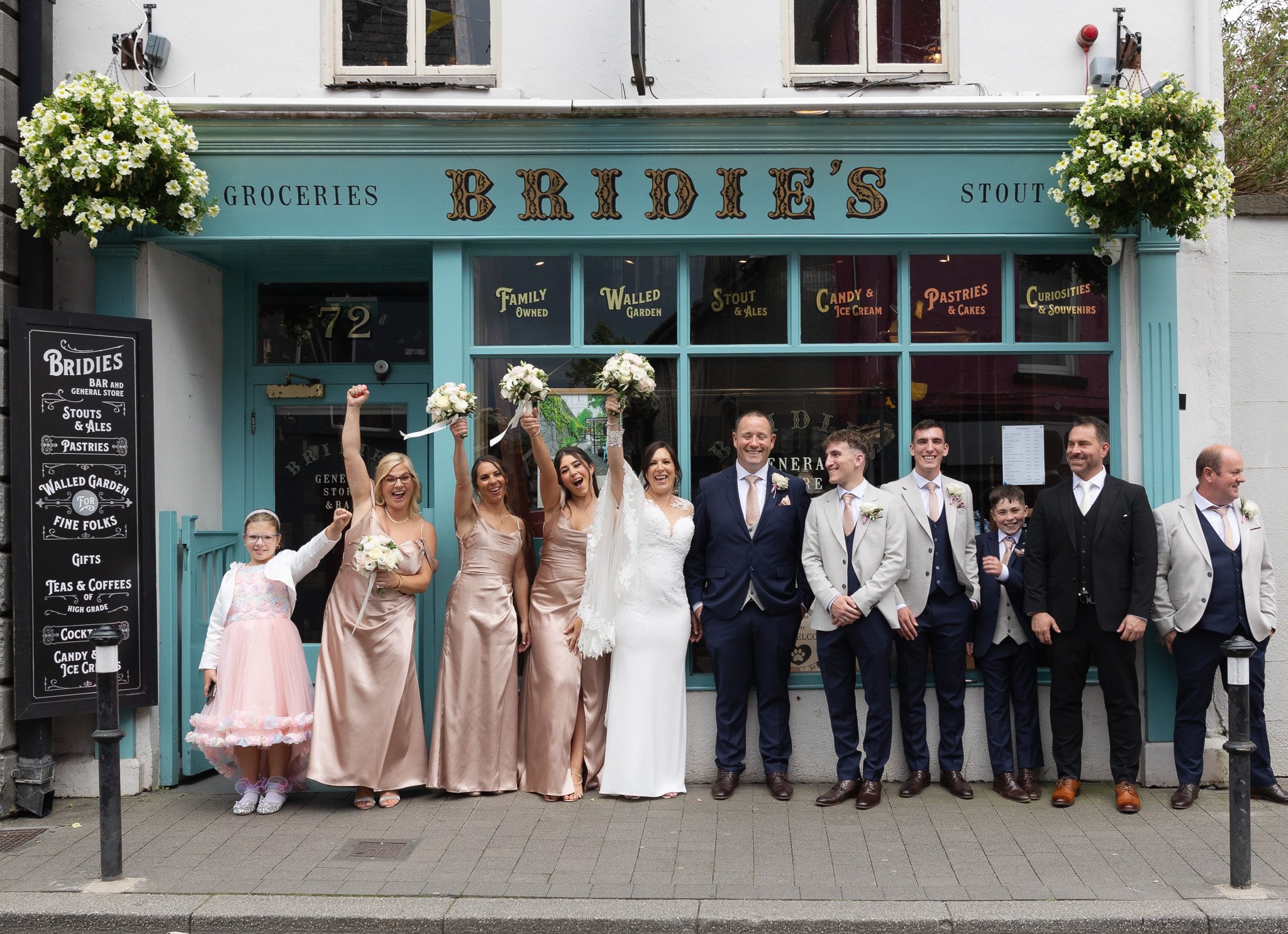 A wedding group photo in front of a store called Briddie's. The group includes a bride in a white wedding dress, a groom in a navy suit, several bridesmaids in matching satin dresses, groomsmen in suits, and a young girl in a pink dress. Some are hol