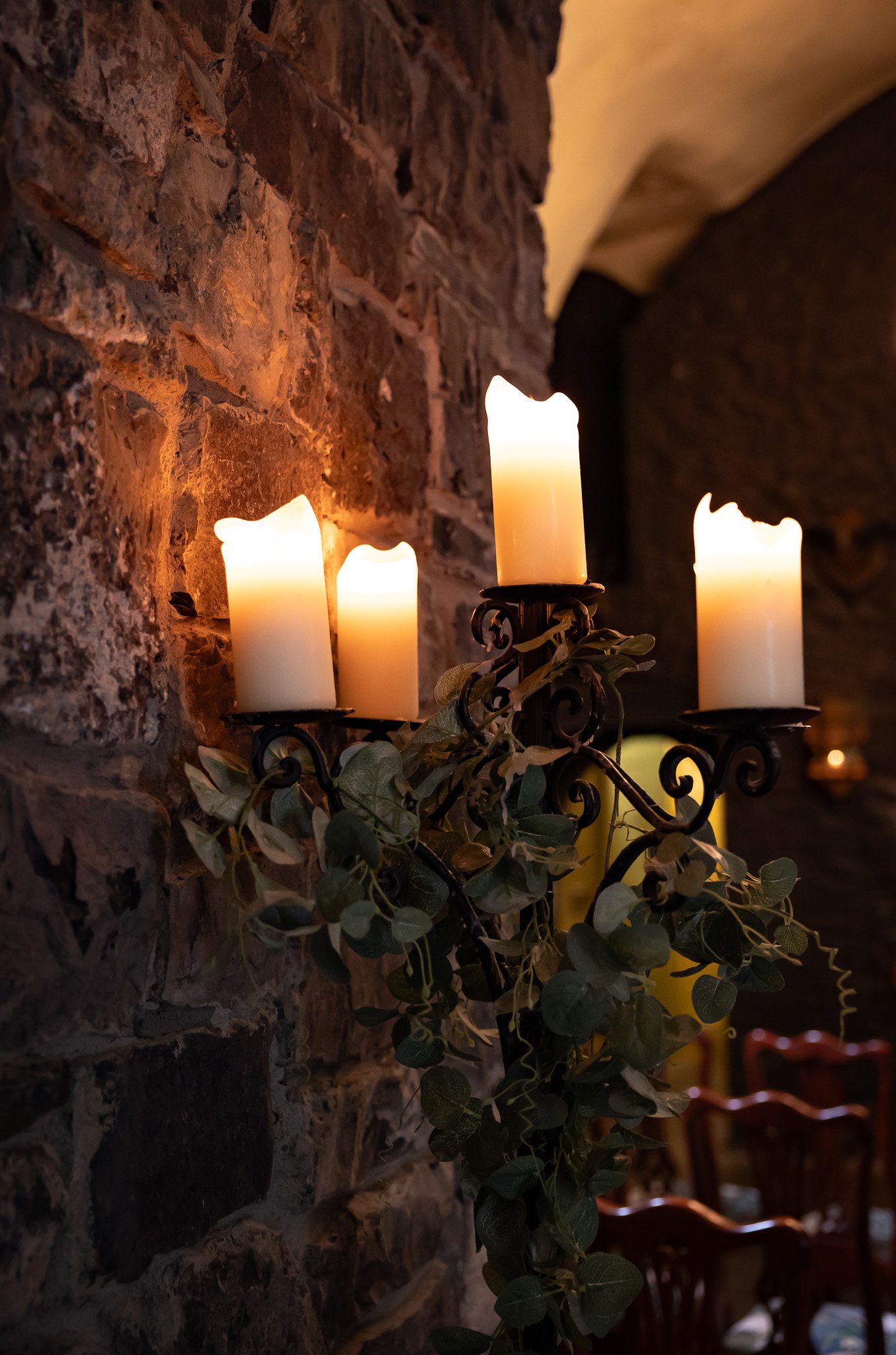 A decorative wall-mounted candle holder with four flickering electric candles, attached to a brick wall, with greenery draped around it, in a dimly lit room.