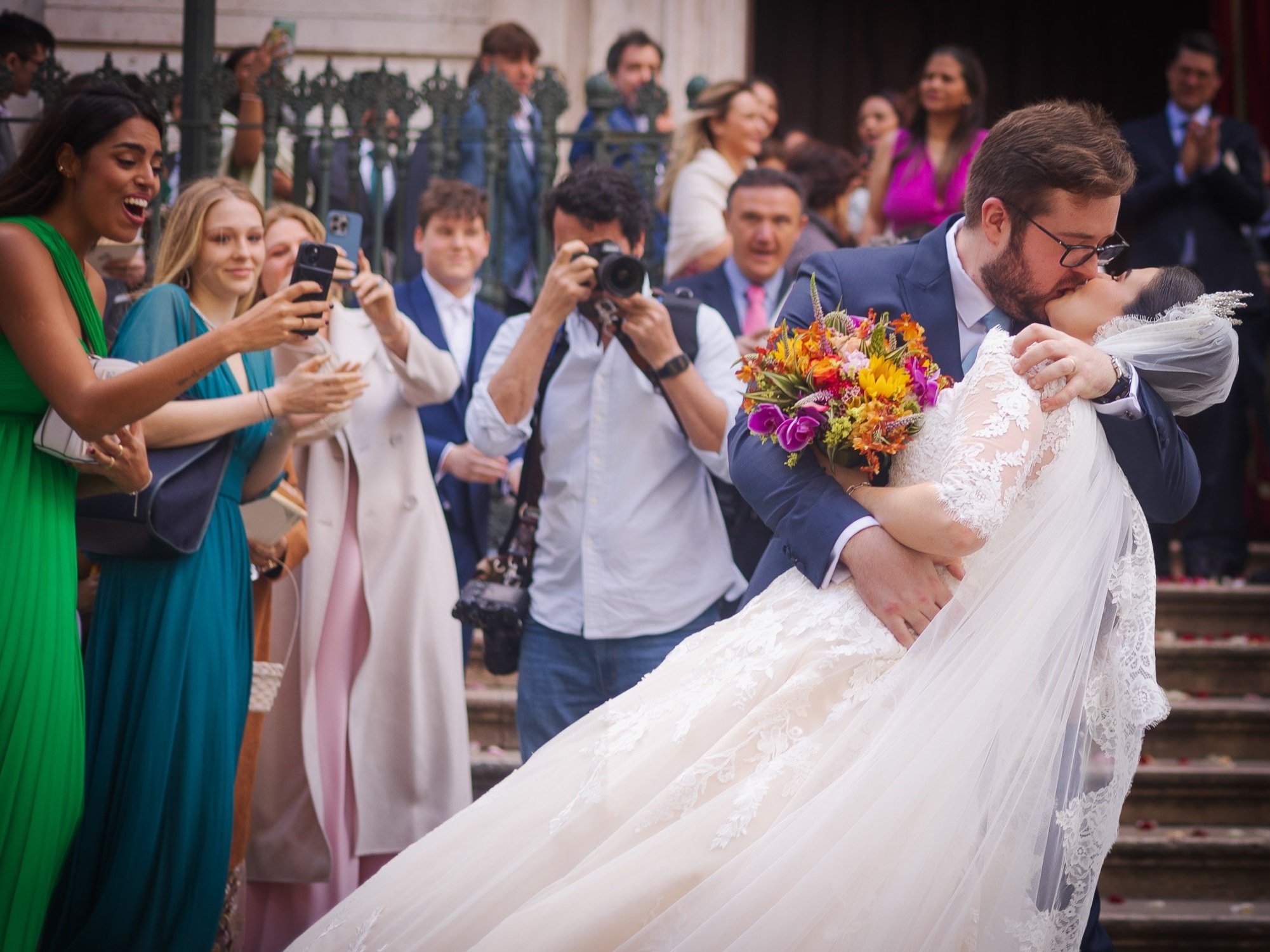 A newlywed couple shares a kiss as the groom dips the bride during their wedding ceremony, surrounded by friends and family. The bride holds a bouquet of colorful flowers, and the guests are capturing the moment with phones and cameras.