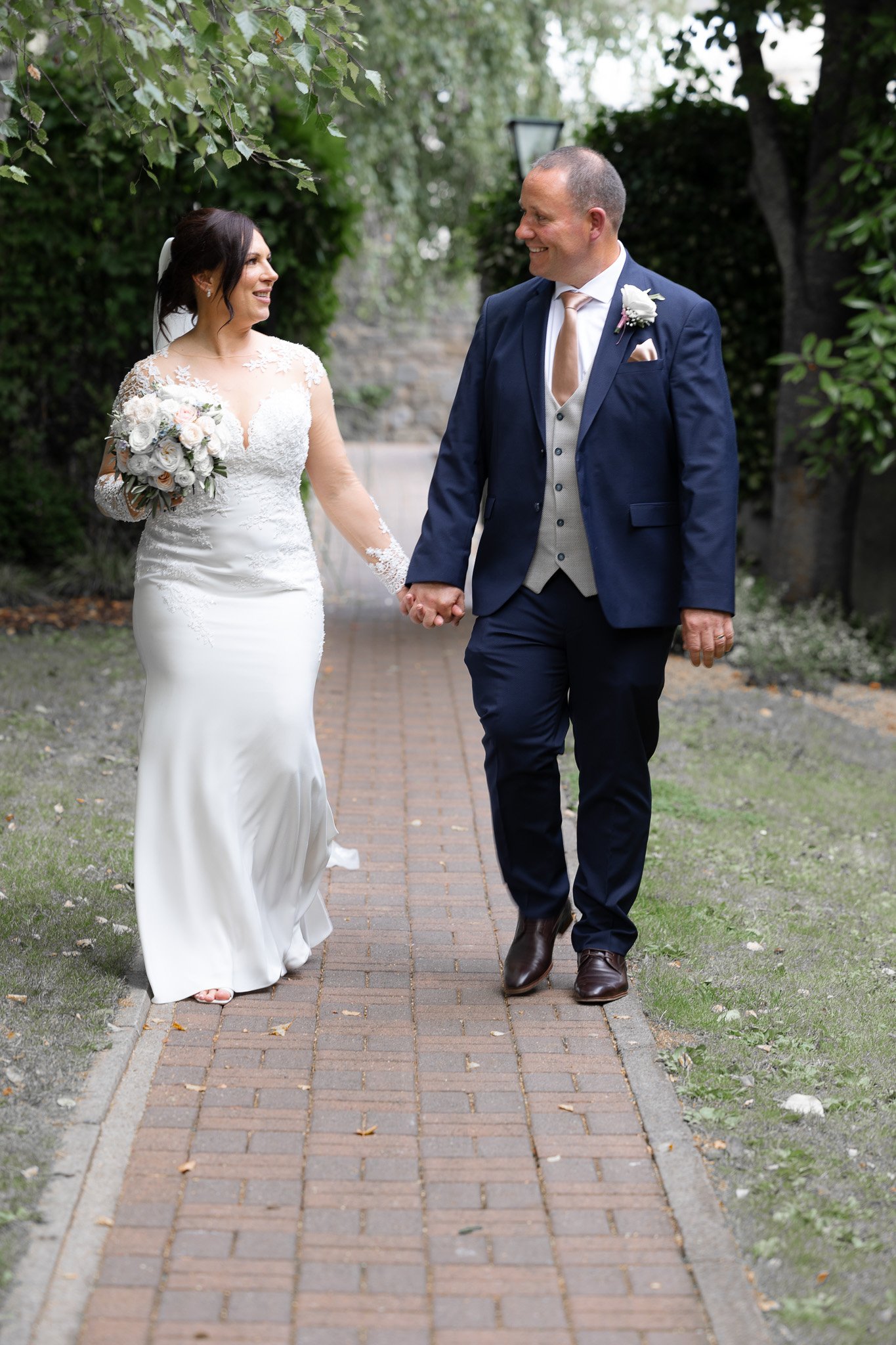 A bride and groom walking hand in hand on a brick pathway outdoors, smiling at each other. The bride is in a white wedding dress holding a bouquet, and the groom is in a navy blue suit with a boutonniere.