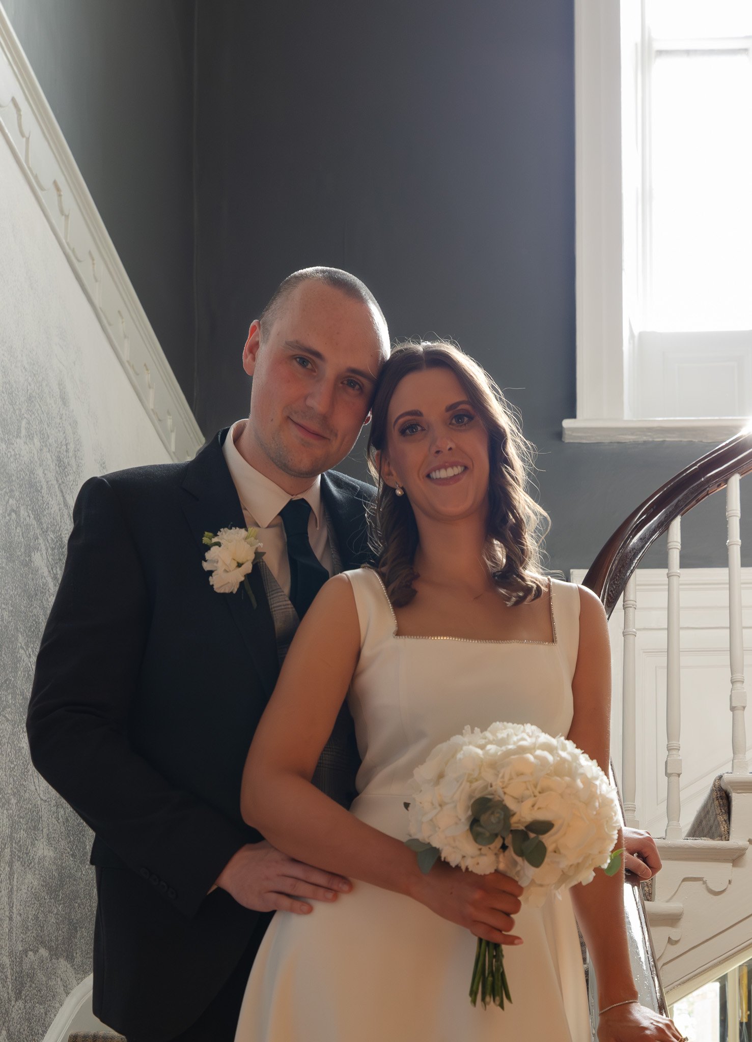 A newlywed couple standing on a staircase indoors, smiling at the camera. The woman is holding a bouquet of white flowers, and the man is dressed in a black suit with a white shirt and a boutonniere. Sunlight is coming through a window behind them.