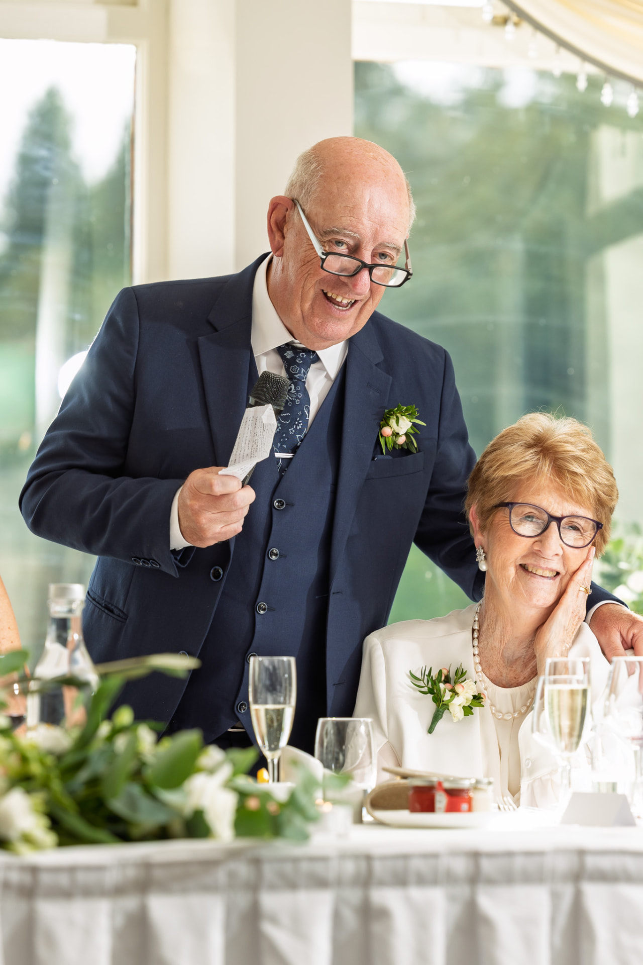 Guest delivering a speech beside a smiling relative at the top table