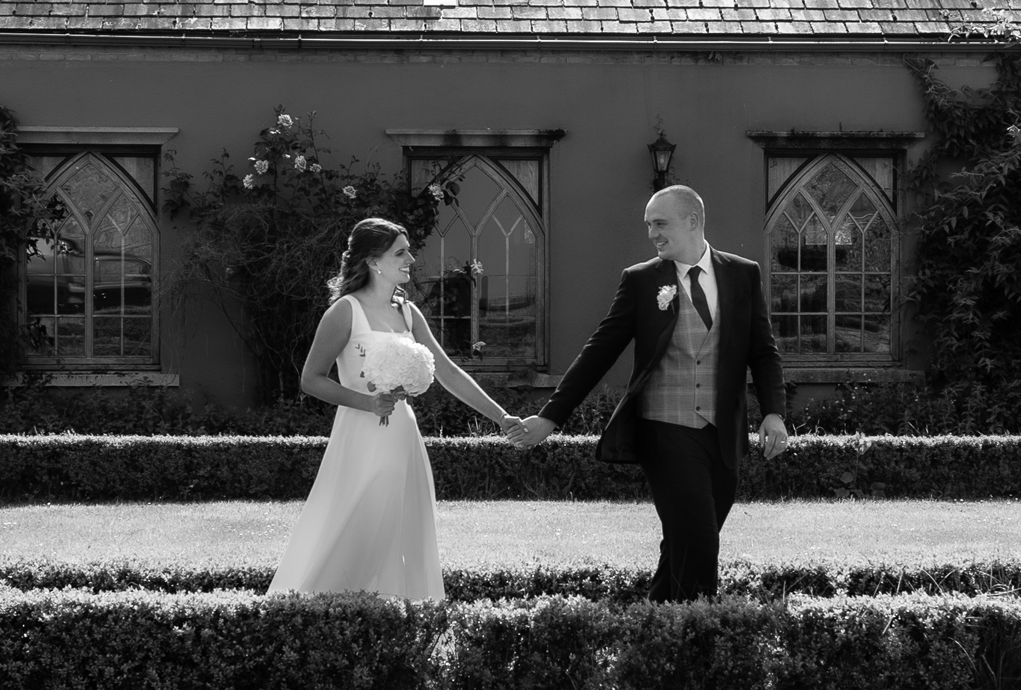 A black and white photo of a bride and groom holding hands and smiling at each other outside, with a background of a building with arched windows and plants.