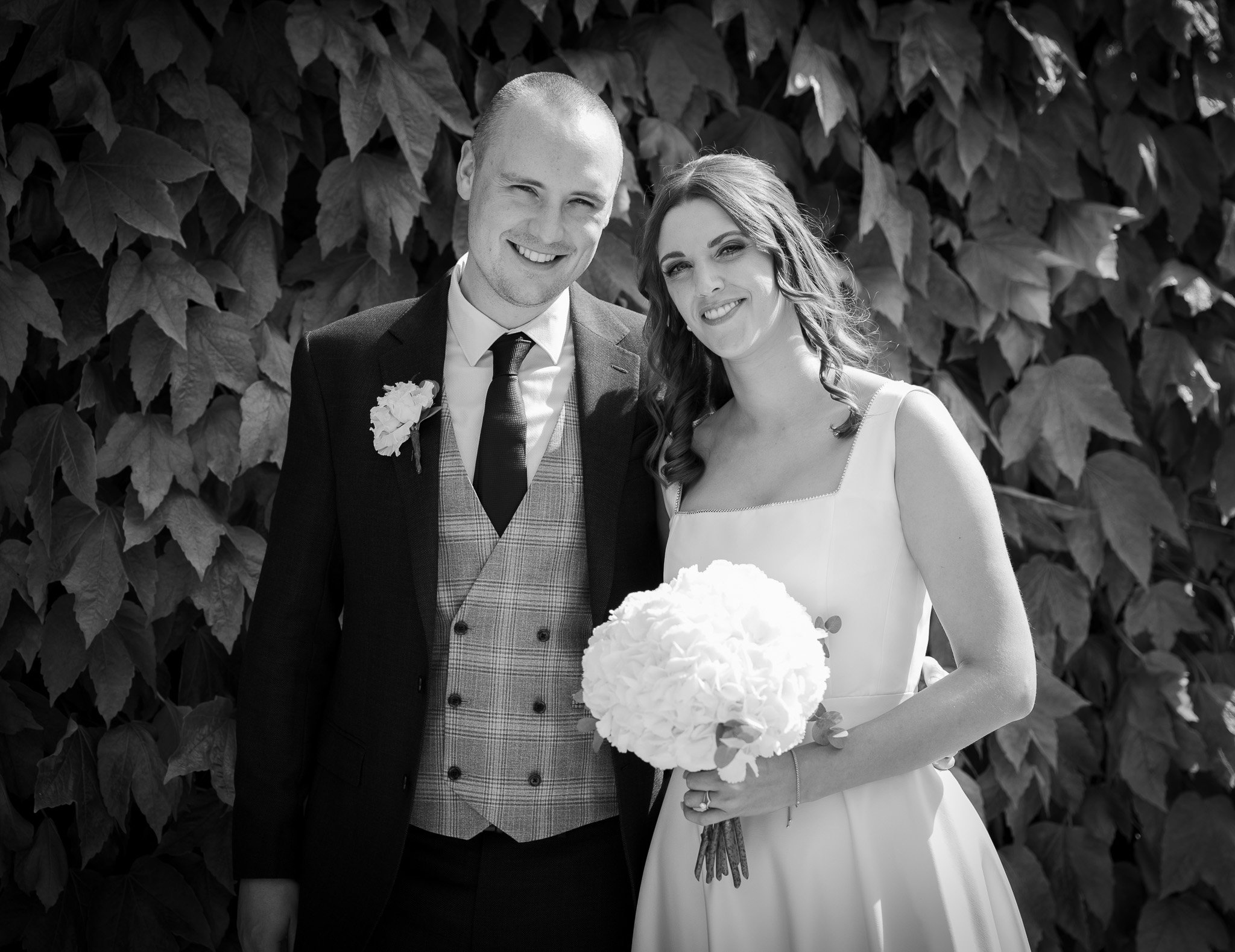 A smiling bride and groom standing close together outdoors in front of leafy background. The groom is dressed in a suit with a plaid waistcoat and boutonniere. The bride is wearing a wedding gown and holding a bouquet of flowers.