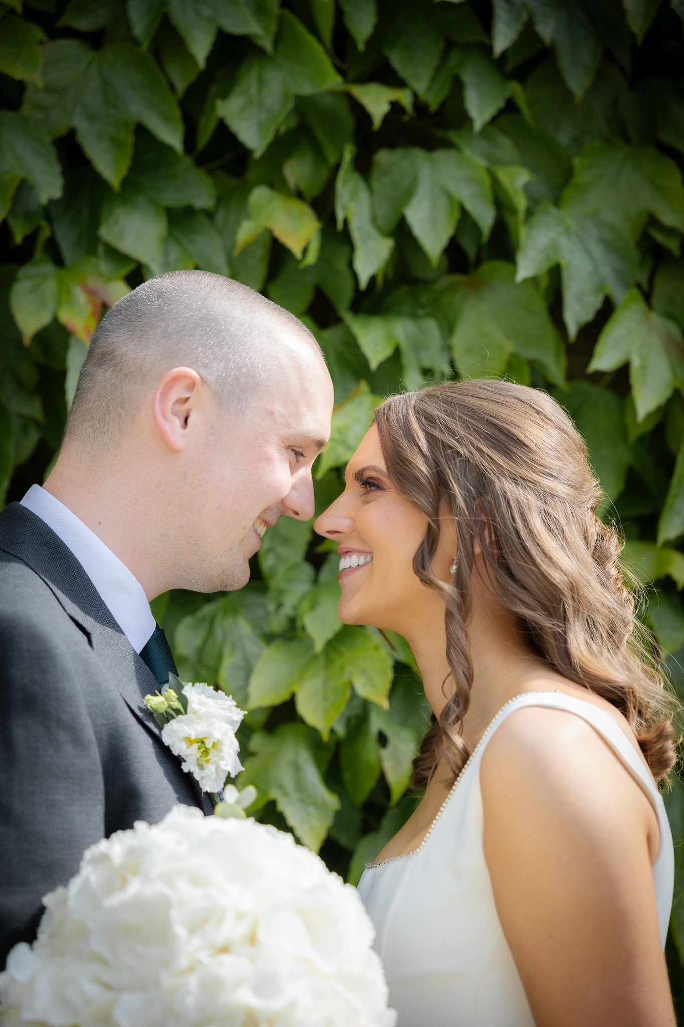A newlywed couple sharing a tender moment, standing close and smiling at each other, with lush green ivy in the background. The bride has long, wavy brown hair and is holding a bouquet of white flowers, while the groom is wearing a dark suit and a wh