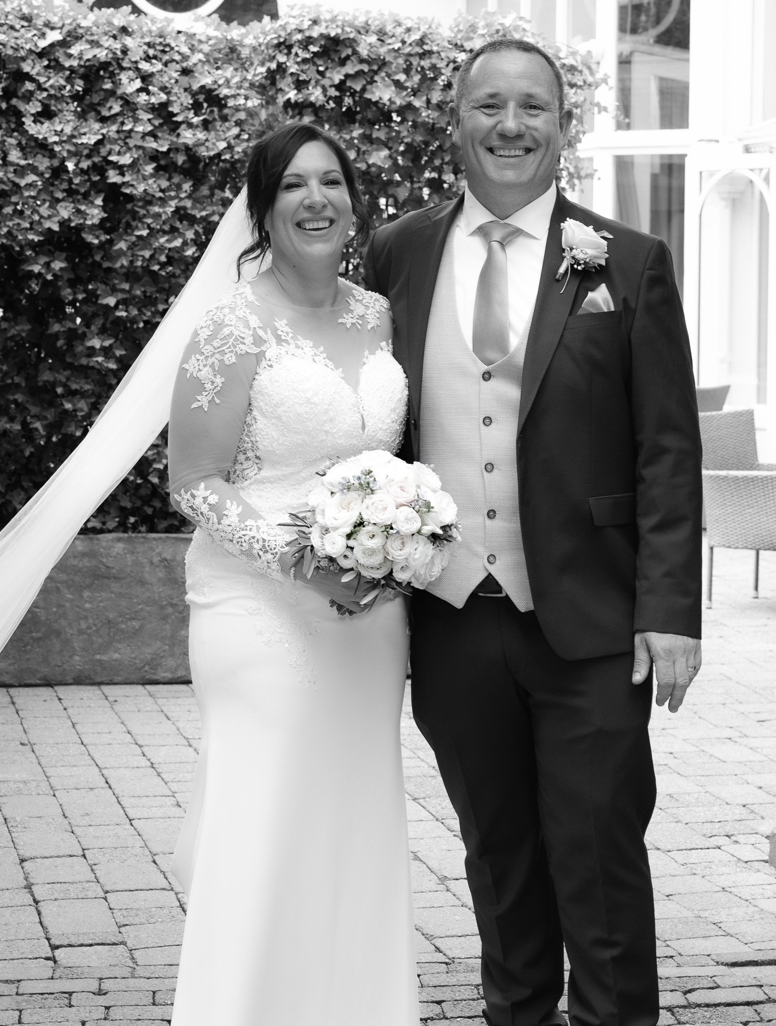 A bride and groom smiling for a photo outdoors on their wedding day, standing on a paved area with a bush and glass building in the background.