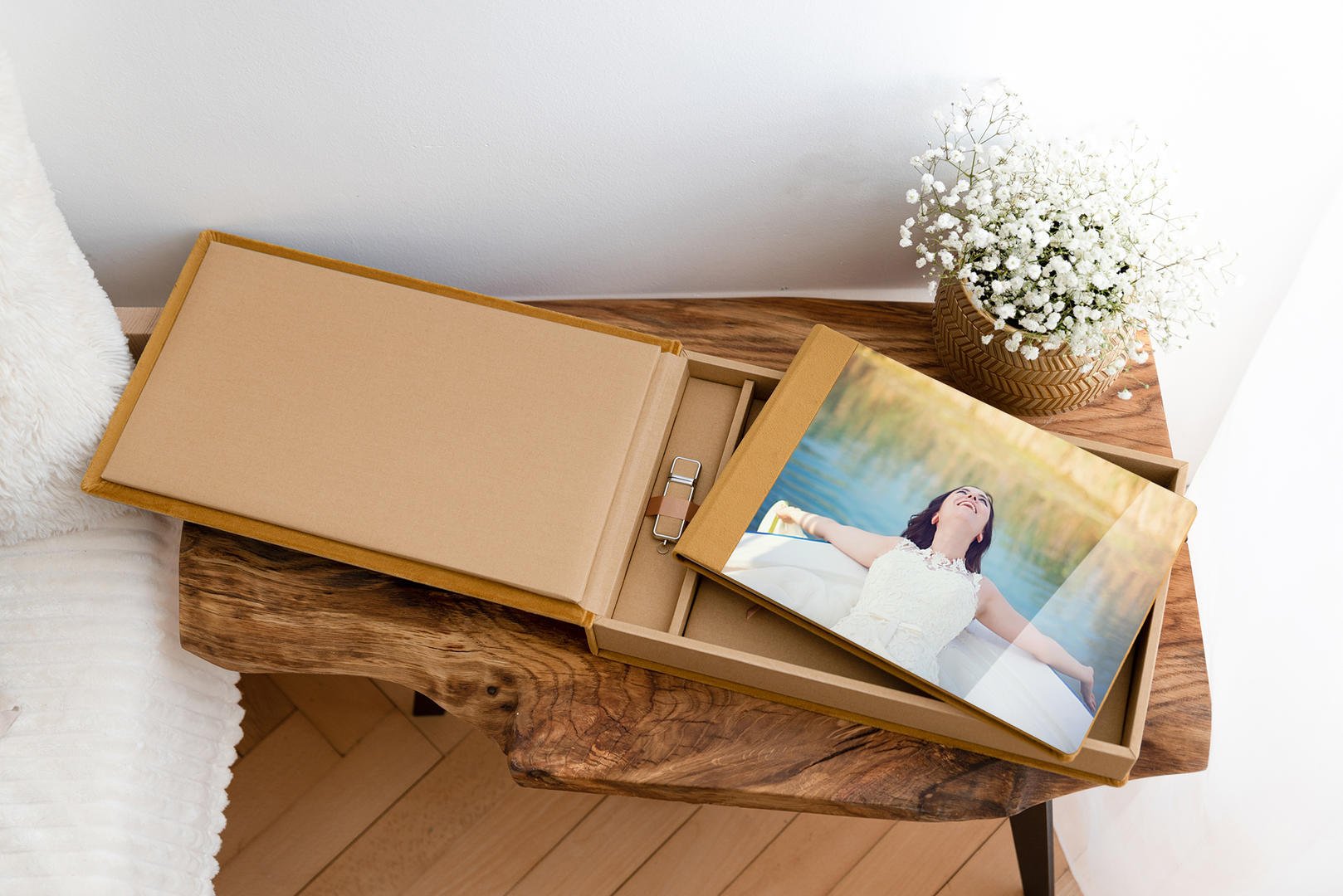 A photo album with a photo of a woman outdoors near a body of water, inside an open cardboard box on a wooden table, next to a basket of white baby's breath flowers.