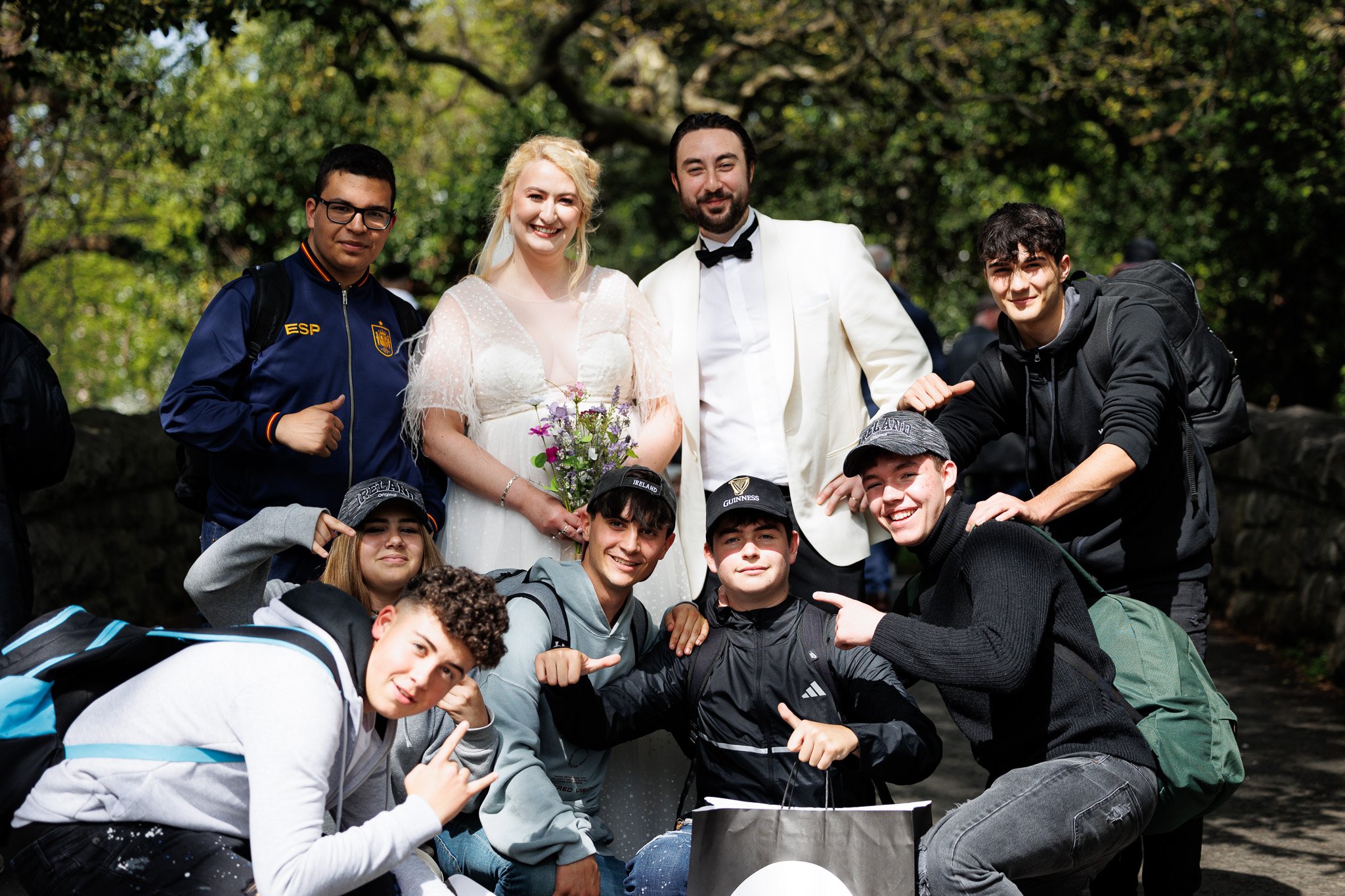 A group of people at a wedding or special event outdoors, including a woman in a white dress holding flowers, a man in a white tuxedo, and several young men in casual and sportswear, smiling and posing for the photo surrounded by trees.
