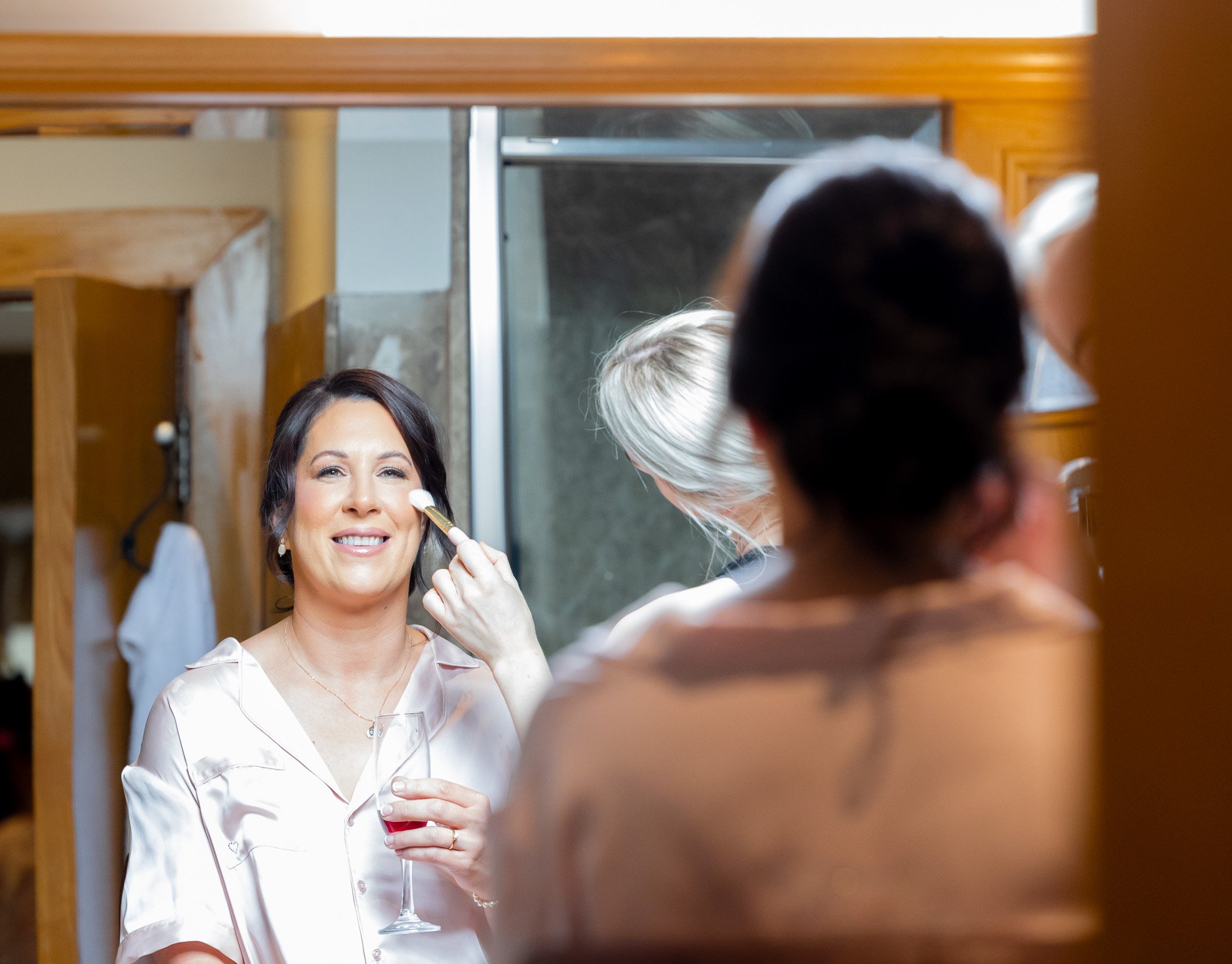 A woman with dark hair getting her makeup done while holding a glass of red wine, with another woman applying makeup in a mirror.