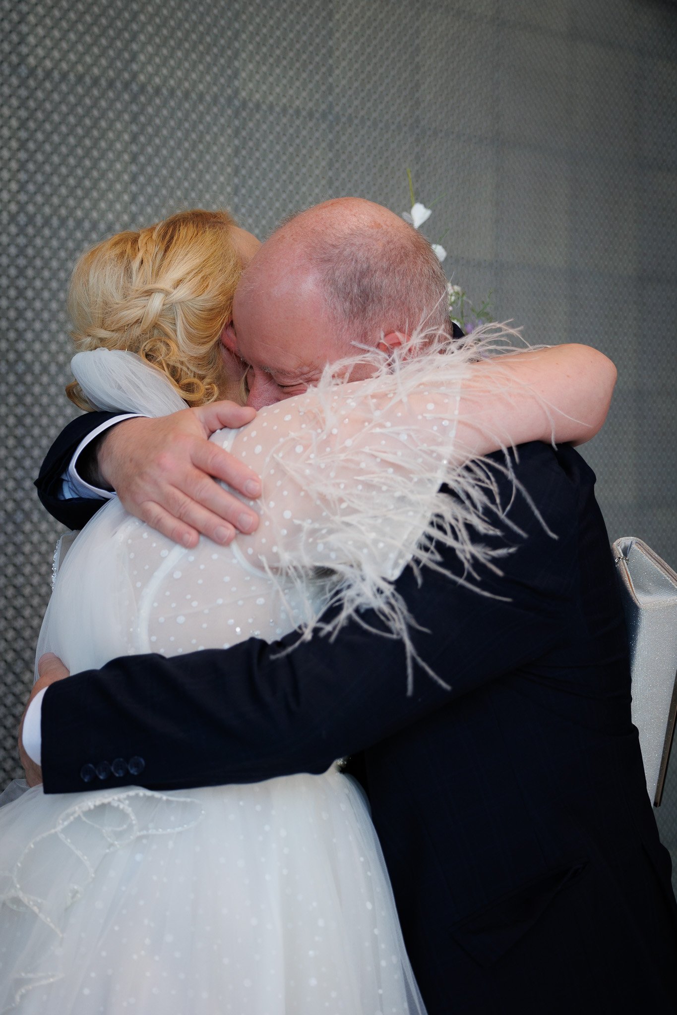 A woman and a man hugging tightly at a wedding, the woman wearing a white dress with feathered details and the man in a black suit.