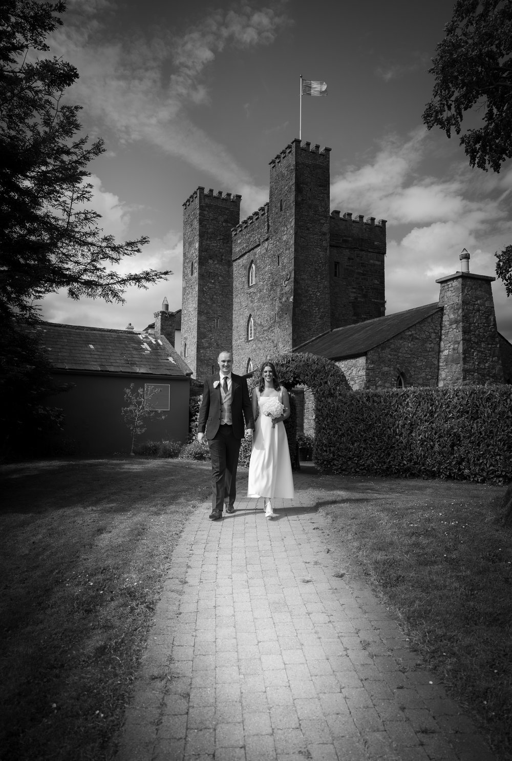 A newlywed couple walking on a brick path in front of a historic castle. The groom wears a suit, and the bride is in a wedding dress holding a bouquet. The scene is outdoors with trees and a partly cloudy sky.