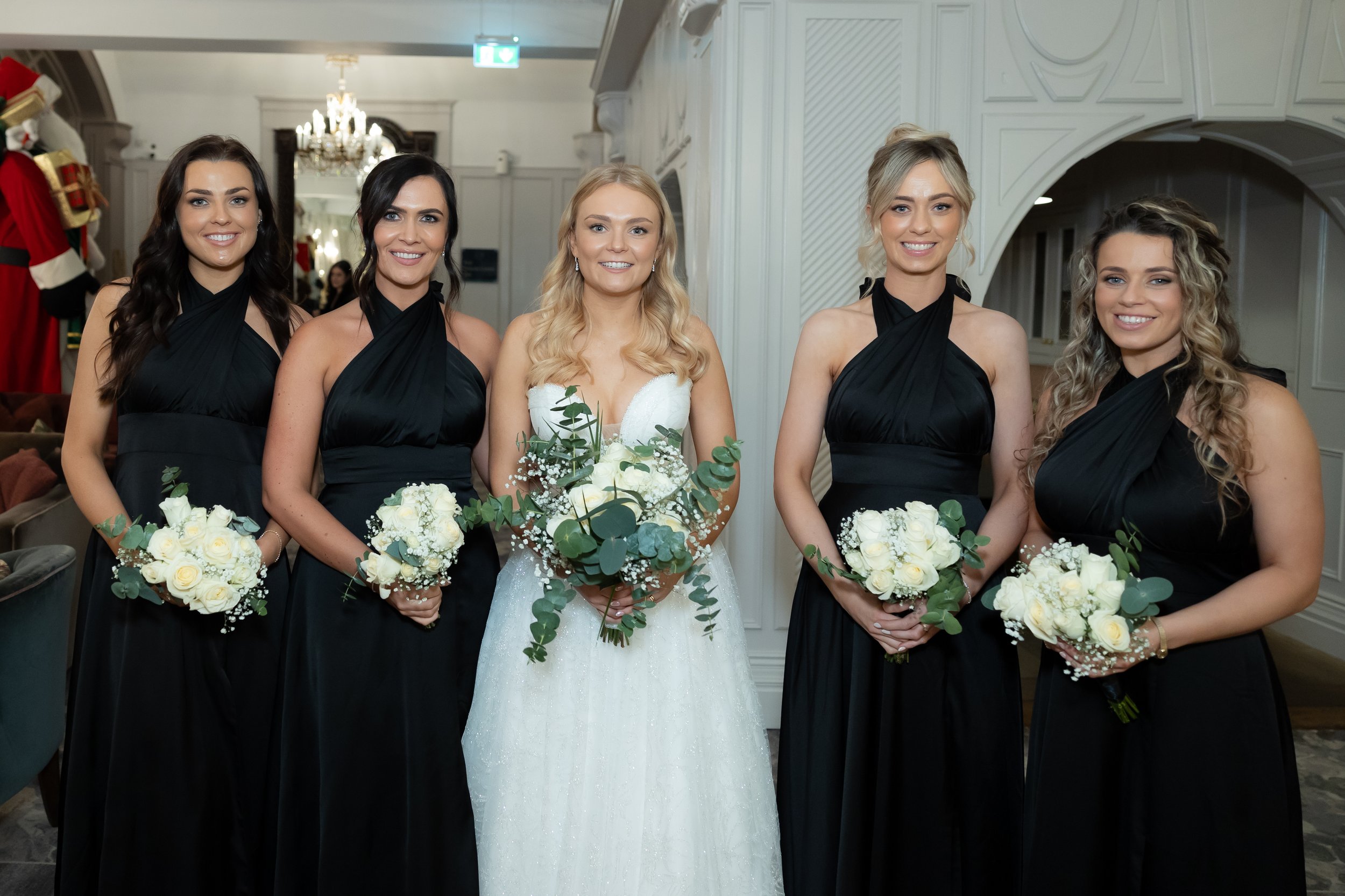 A bride in a white wedding dress with long blonde hair stands between four women in black bridesmaid dresses, all holding white floral bouquets, inside a decorated room.