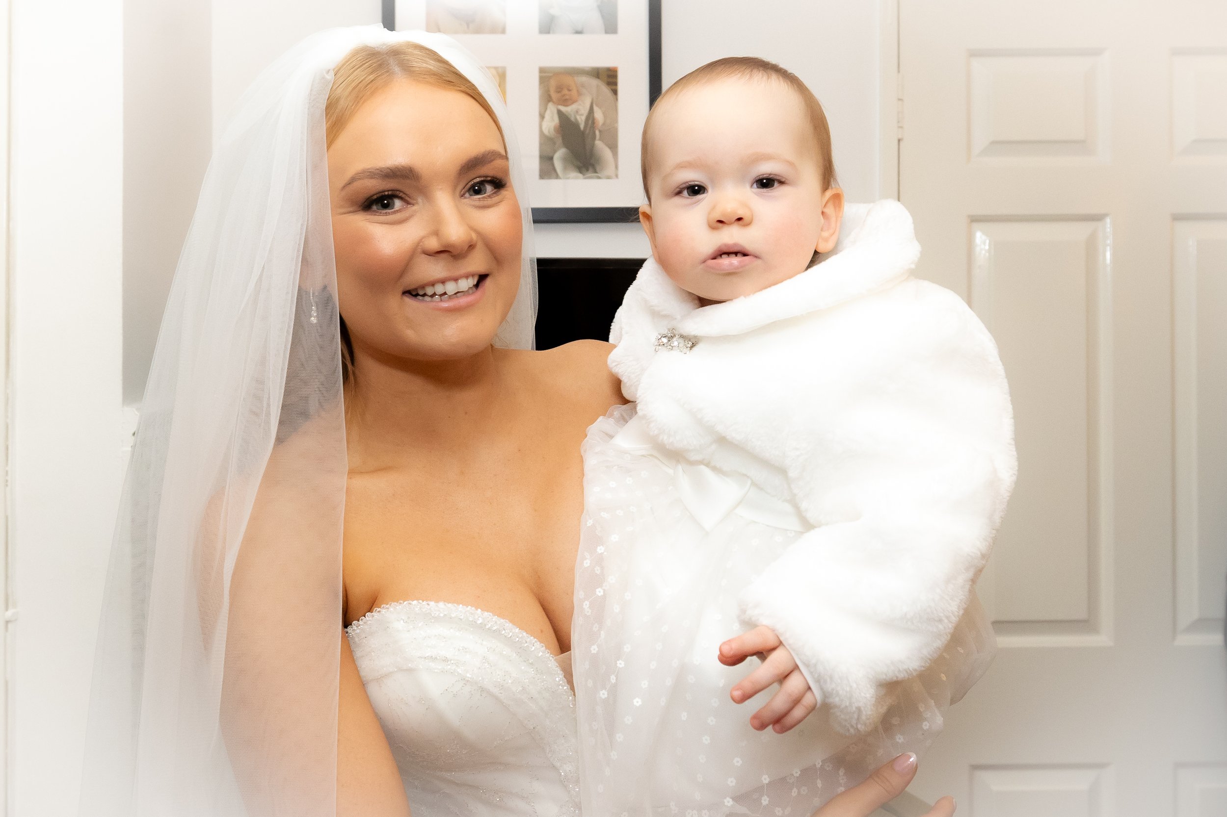A smiling woman dressed as a bride holding a young child dressed in white, in front of a white door with framed photos on the wall behind them.
